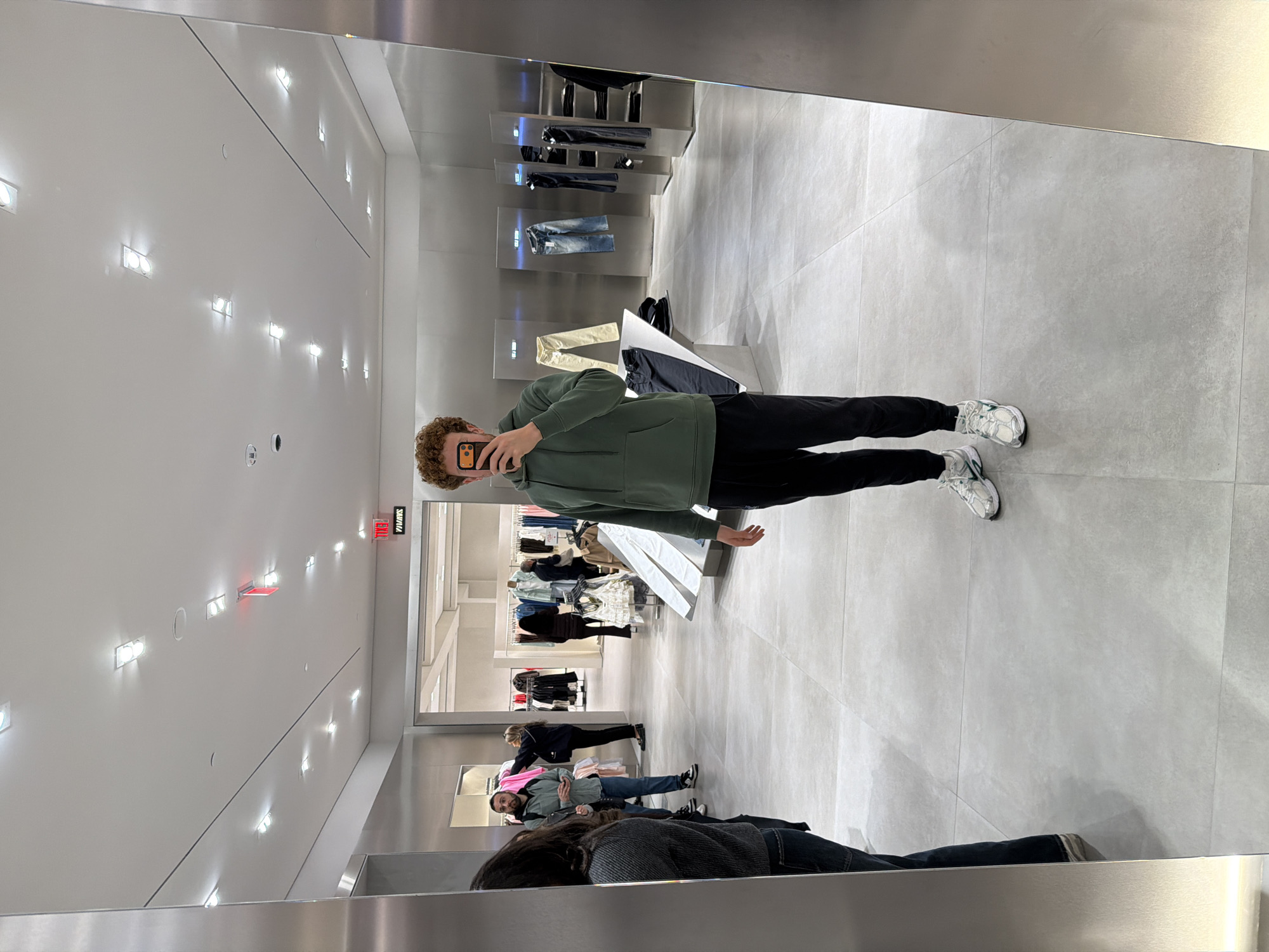 A photograph taken in a store in New York City, United States of America. The image appears to be a selfie taken using a reflective surface such as a mirror or a glass window.
In the foreground, a person is holding a phone taking the picture. This individual is wearing a green hoodie and black pants, along with white sneakers. Behind the person, in what appears to be the main store area, are clothing racks and displays with various garments. Several other people are browsing or walking around in the store. The store has a modern and minimalist aesthetic with light-colored flooring and bright lighting. The environment suggests a retail setting, likely a clothing store.