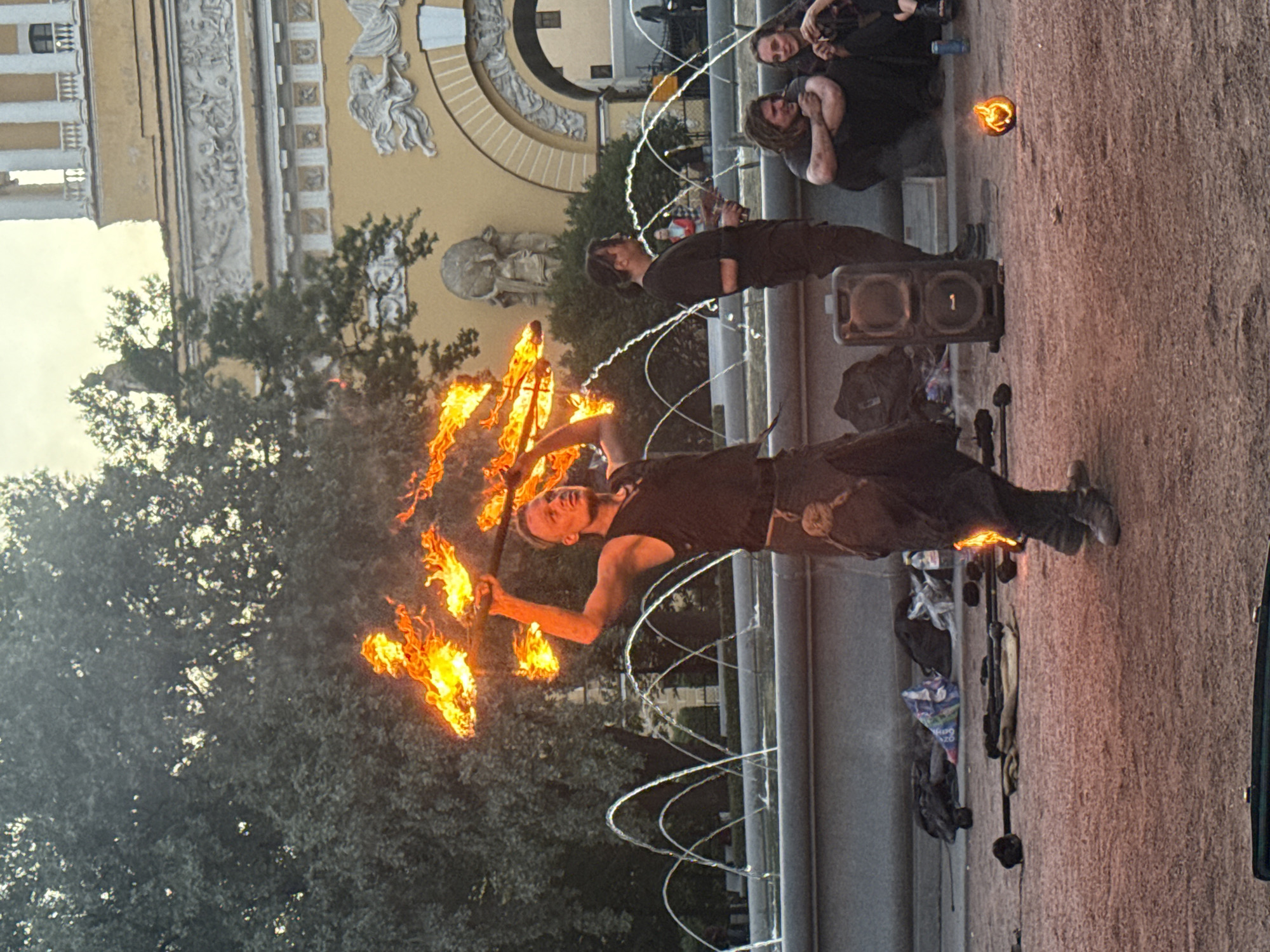 A male fire performer is the central subject, wielding a flaming prop above his head. He is dressed in dark clothing, including a vest and trousers, and appears to be in the midst of a performance. In the background, a fountain with multiple arcs of water is visible, and behind that, a large, ornate yellow building with classical architectural elements stands. Several other people are present, some seated and observing the performance, while others stand. One person, also dressed in dark clothing, stands to the right of the fire performer, holding a drink. To the far right, two individuals are seated on the ground, and a small ball of fire is visible on the ground in the foreground. The ground appears to be a reddish-brown gravel or dirt surface. Various props, including what seem to be unlit fire-staffs, are scattered on the ground around the performers. The overall scene suggests an outdoor performance or event taking place in an urban plaza. The architecture of the building in the background strongly suggests the location is Palace Square, Saint Petersburg, Russia, likely in front of the Winter Palace or the General Staff Building.