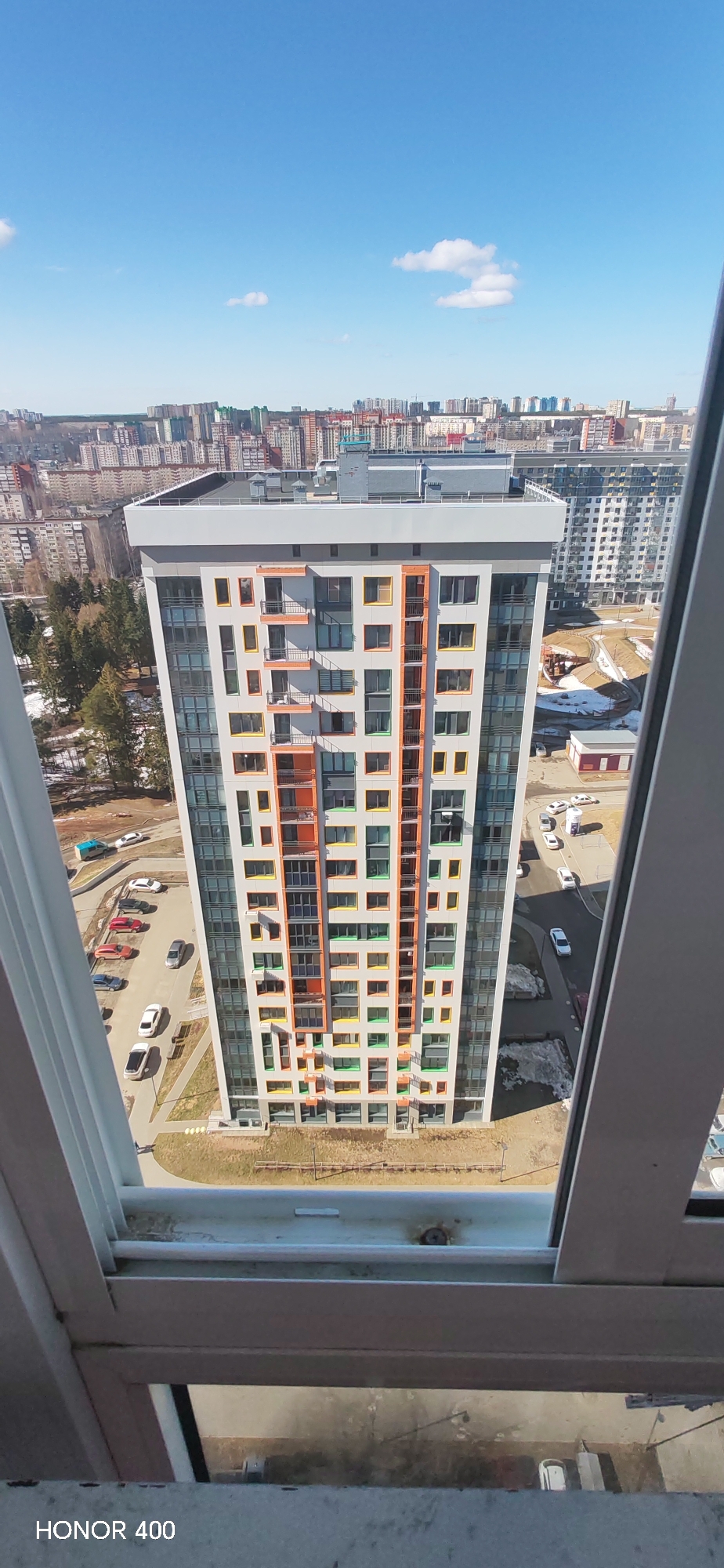 The image is a high-angle view from a window, looking out at a modern building in Izhevsk, Russia. The building has a rectangular footprint and multiple stories, likely more than 20, with a flat roof. The exterior is characterized by a repetitive pattern of windows framed in various colors like orange, yellow, and green, contrasting with the gray or white building facade. The architectural style appears contemporary, with a mix of large glass panels and solid wall sections.
In the background, a cityscape unfolds with numerous other buildings of varying heights and architectural styles, indicating a dense urban environment. A patch of trees is visible in the middle ground, suggesting a park or green space within the city.
Below the building, there is evidence of a parking lot with cars, and a paved area, suggesting vehicle and pedestrian traffic in the vicinity. The day is sunny, with a clear blue sky punctuated by a few scattered clouds, enhancing the visibility and sharpness of the scene.
The perspective suggests the photograph was taken from a high vantage point, possibly within another building. The lower portion of the frame includes part of the window frame, providing context for the viewpoint. A watermark or branding "HONOR 400" is visible at the bottom left of the image.
