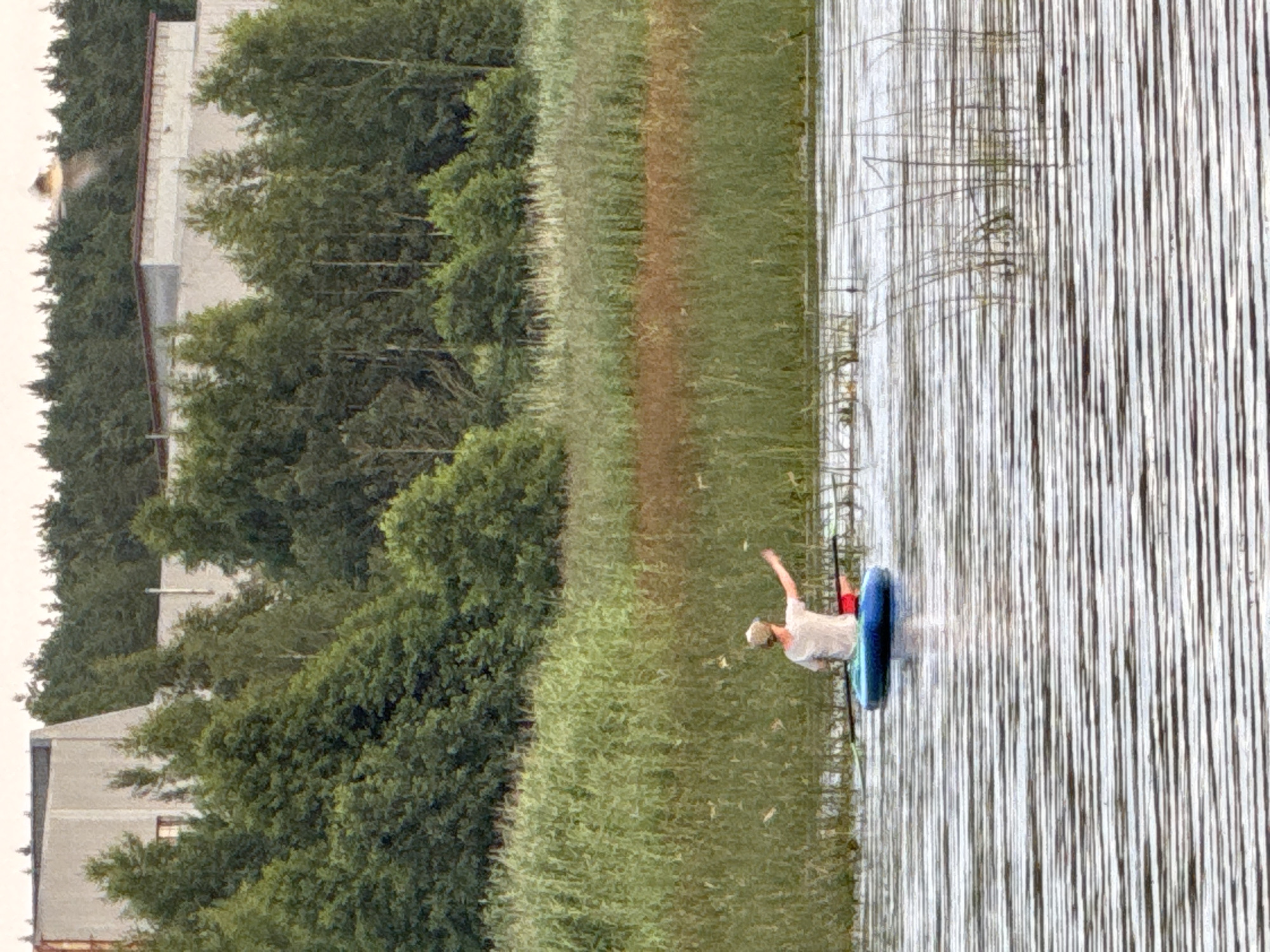 In Glebychevo, Russia, on a body of water, a person is seated in a blue kayak. The individual is wearing a light-colored t-shirt and a hat, with their right arm extended. The kayak is positioned in front of a dense, tall grass shoreline, which is bordered by a line of trees. In the background, there are two large, light-colored industrial buildings visible above the trees. The water is rippled, reflecting the overcast sky.