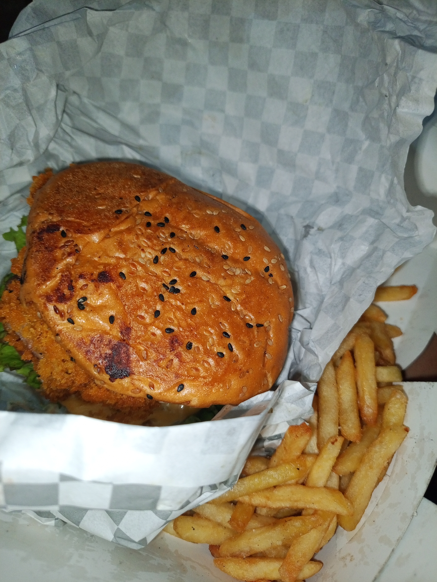 A high-angle, medium shot shows a burger and fries in a white paper container with a gray and white checkered pattern, likely a takeout container. The burger has a browned, sesame-seed-covered bun, visible breaded patty, and some green lettuce peeking out. To the right of the burger are golden-brown French fries. The photograph was taken indoors. Tinaquillo, Venezuela is not evident in the image.
