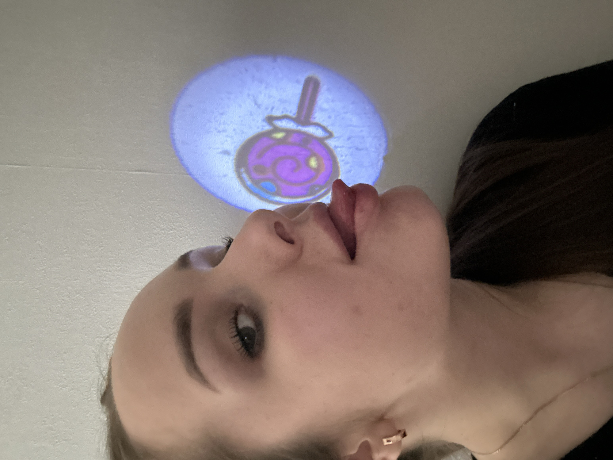 A close-up, low-angle shot captures a young person, likely female, in an indoor setting, possibly in Chaikovsky, Russia. The individual is positioned on the left side of the frame, looking upwards with her head tilted back and her tongue slightly extended. Her gaze and tongue are directed towards a circular, luminous projection displayed on a light-colored, textured surface, presumably a ceiling or wall, above her.
The projection vividly depicts a cartoon-style purple lollipop or candy, featuring a swirling design, a stick, and smaller yellow and blue accents. The lighting in the room appears dim, allowing the bright, glowing projection to stand out.
The person has fair skin, visible eyelashes, and dark hair that falls over her shoulder. She is wearing a small, golden hoop earring in her right ear and a thin, delicate necklace around her neck. Her expression seems playful and engaged, suggesting a lighthearted interaction with the projected image. No text is visible in the image.