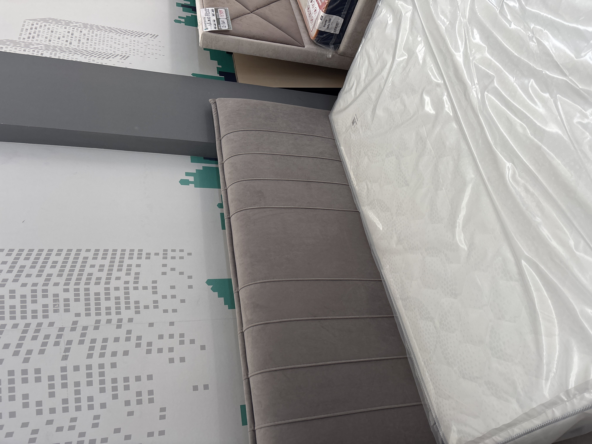 A soft grey upholstered headboard of a bed is visible in the foreground, with a white mattress covered in clear plastic sheeting. Behind the bed, a grey cylindrical pillar stands against a white wall with a graphic design of grey building silhouettes. To the right, another bed with a similarly upholstered headboard, this one featuring a diamond pattern, is visible. A price tag with Cyrillic text and numbers is attached to the second bed. The overall scene suggests a furniture store in Zaporizhzhia, Ukraine.