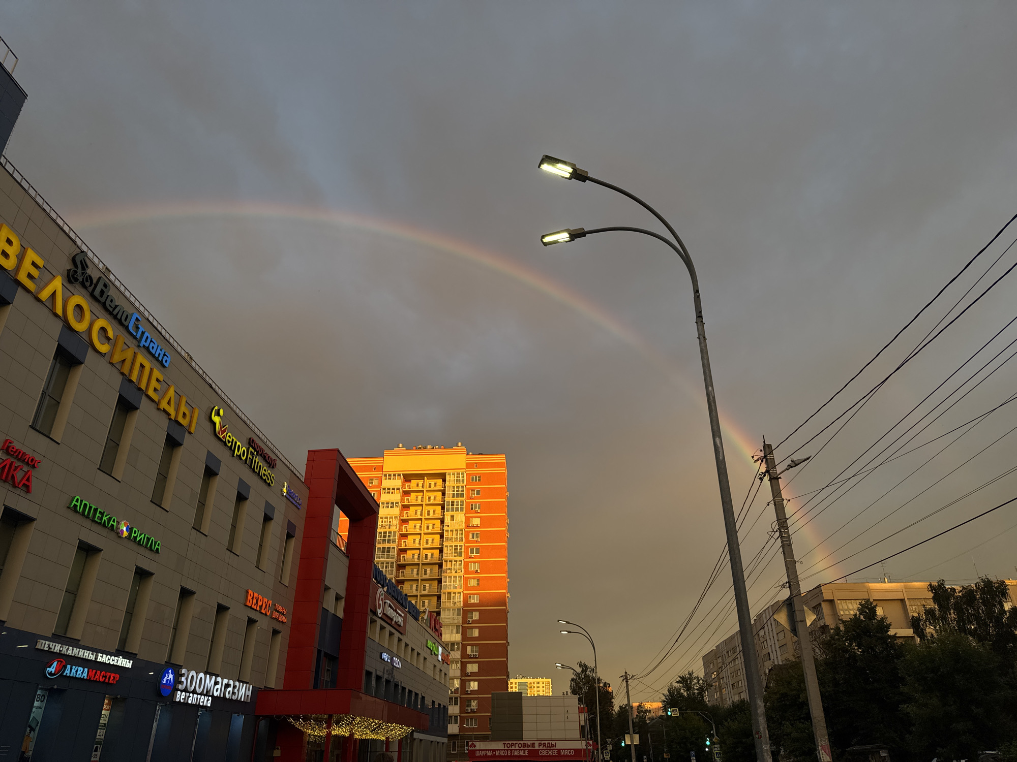 This image captures an urban street scene in Nizhny Novgorod, Russia, under a dramatic sky. A vibrant rainbow arcs across the overcast, grey clouds, suggesting recent rain or ongoing showers transitioning to clearer skies. The low angle of the sun, indicated by the warm, golden light illuminating the side of a tall, orange apartment building, points to either sunrise or sunset.
The foreground features the facade of a multi-story building with various shops and businesses. Prominent signage in Russian includes "ВелоСтрана" (VeloStrana - Bicycle Country), "Metro Fitness," "АПТЕКА РИГЛА" (Apteka Rigla - Rigla Pharmacy), "АКВАМАСТЕР" (Akvamaster), and "Зоомагазин Ветаптека" (Zoomagazin Vetapteka - Pet Store Veterinarian Pharmacy). Another sign reads "ВЕРЕС ТОВАРЫ ДЛЯ ДОМА" (Veres Tovary Dlya Doma - Veres Home Goods). Further down the street, a sign for "ТОРГОВЫЕ РЯДЫ" (Torgovye Ryady - Market Rows) advertises "ШАУРМА МЯСО В ЛАВАШЕ СВЕЖЕЕ МЯСО" (Shaurma Myaso v Lavashye Svezheye Myaso - Shawarma Meat in Lavash Fresh Meat).
Two streetlights with bright LED bulbs are visible in the mid-ground, their illumination cutting through the subdued natural light. Electrical wires crisscross the sky, a common sight in urban environments. The overall atmosphere is one of urban life continuing despite, or perhaps enhanced by, the natural phenomenon of the rainbow and the atmospheric lighting. The scene evokes a sense of normalcy and resilience in a city setting.