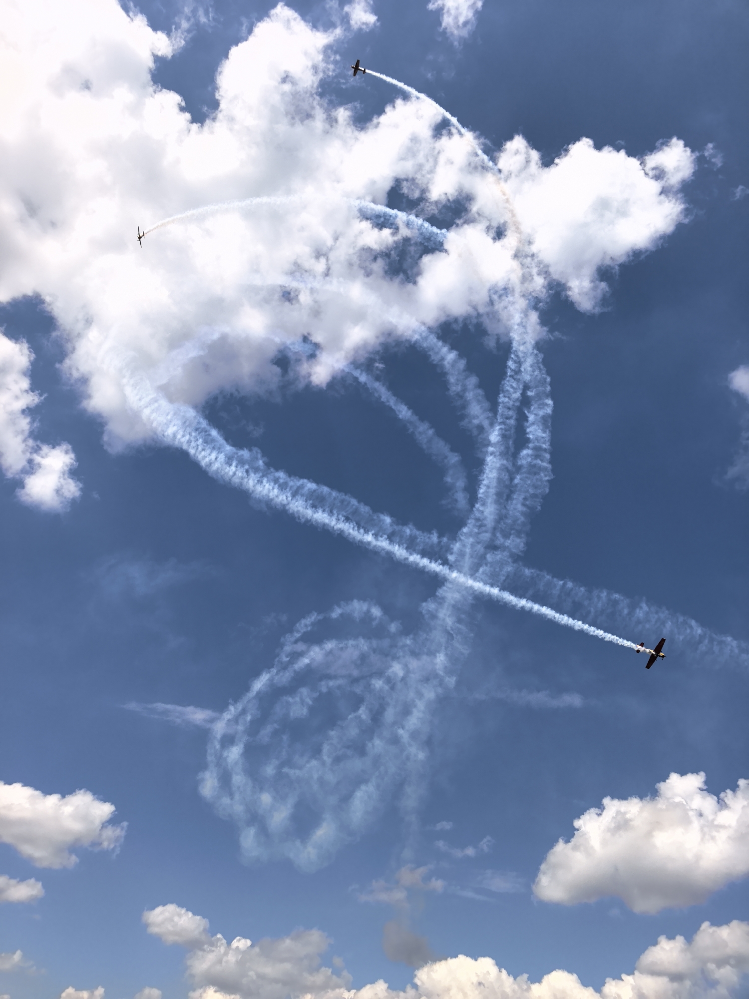 This image depicts an air show with three small airplanes performing aerial maneuvers, leaving behind white smoke trails against a bright blue sky with scattered white clouds. The airplanes are small, single-engine propeller aircraft. One airplane is visible in the upper right quadrant, trailing smoke. Another is in the upper left quadrant, also trailing smoke. A third airplane is in the lower right quadrant, moving diagonally upwards and to the left, leaving a long, distinct smoke trail. The smoke trails form various patterns, including loops and straight lines, suggesting synchronized or individual aerobatic performances. The scene is likely taking place outdoors during daylight, under a clear or partly cloudy sky. The image does not provide any information or visual cues to identify the specific location as Nestiukovo, Russia.
