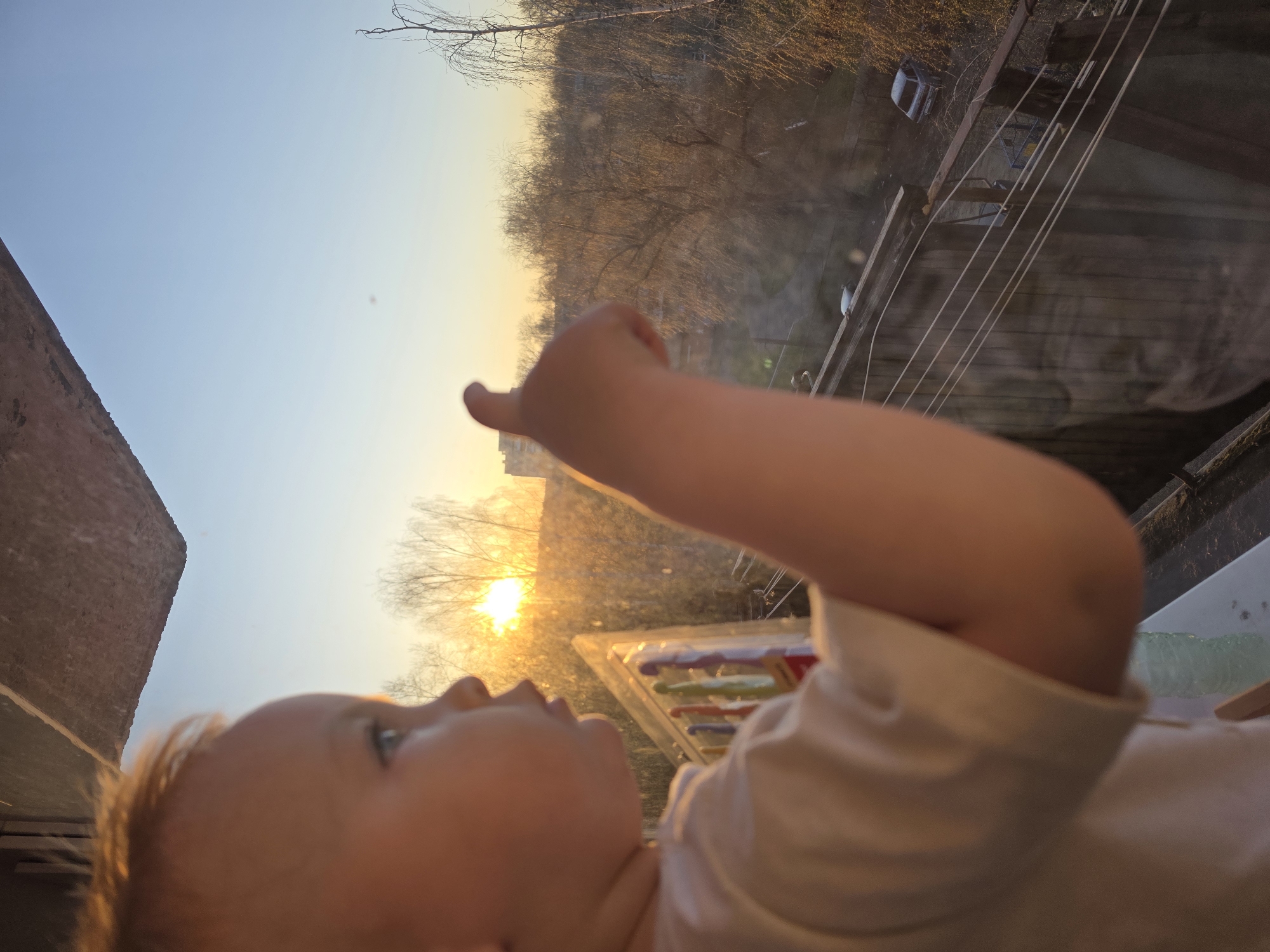 A child in a white shirt lies on their back indoors, facing upward and pointing with their right index finger towards a sunset seen through a window. The child appears to be near a window that is reflective, showing bare trees and a building across the way. Outside, there are signs of a building and a parking lot with vehicles and power lines. While not explicitly visible, the location is presumably within the city of Chaikovsky, Russia, based on the user's request.