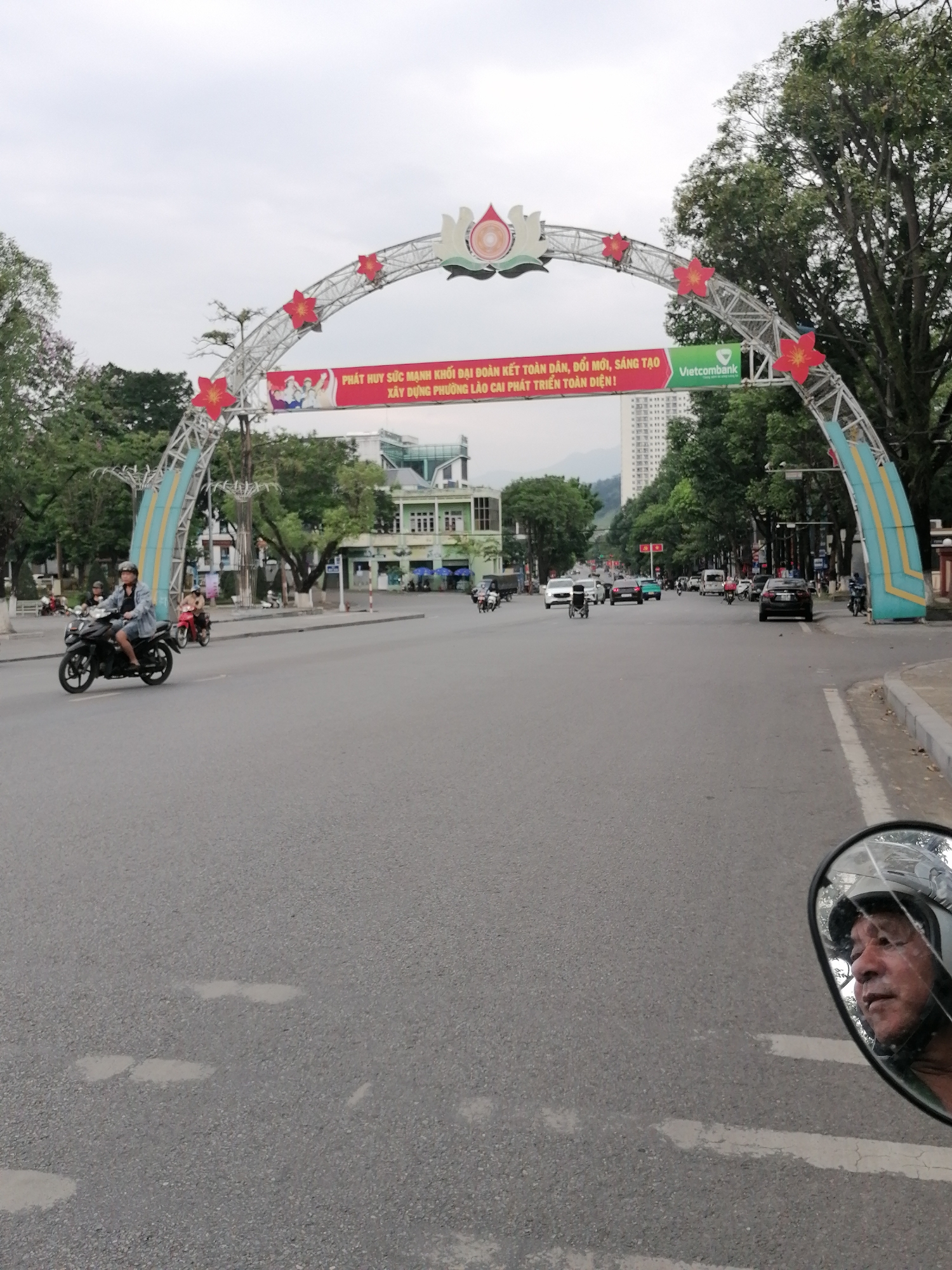 A person is riding a black motorcycle on a wide street in Lao Cai Ward, Vietnam. An arch spans the street. The arch has red flowers, a lotus symbol at the top, and a banner that reads "PHÁT HUY SỨC MẠNH KHỐI ĐẠI ĐOÀN KẾT TOÀN DÂN, ĐỔI MỚI, SÁNG TẠO XÂY DỰNG PHƯỜNG LÀO CAI PHÁT TRIỂN TOÀN DIỆN!" and "Vietcombank". Other vehicles are on the street. Buildings and trees line the street. The motorcyclist's face is visible in the motorcycle's mirror.