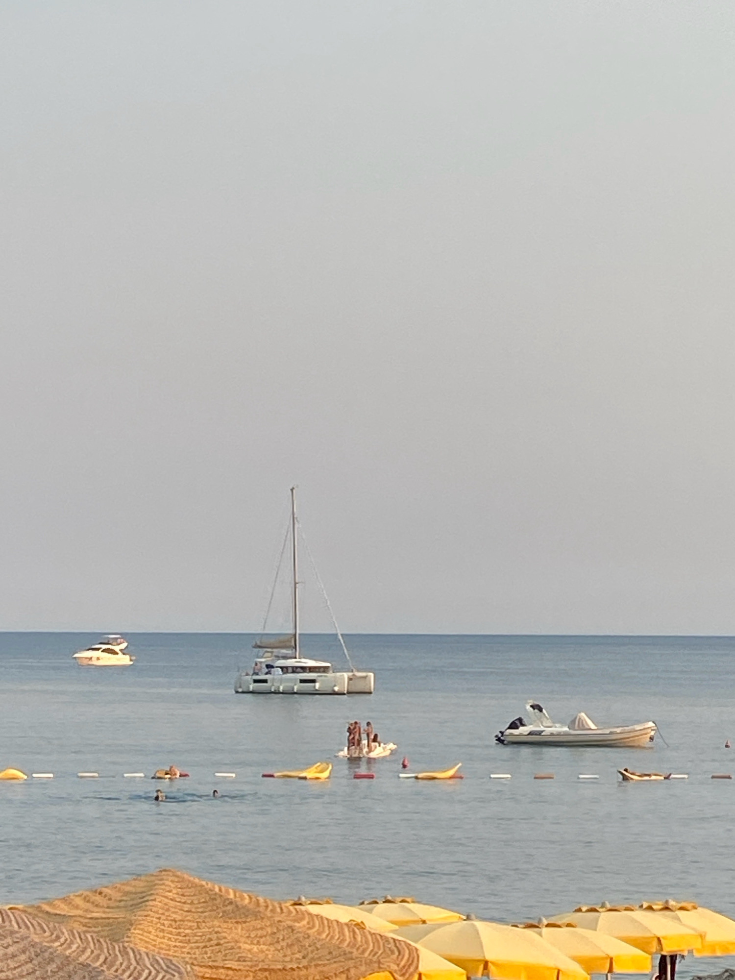 In the sea off the coast of Prijevor, Montenegro, a serene scene unfolds. The calm, blue water stretches towards a hazy horizon, meeting a pale, overcast sky. Several vessels dot the expanse: a sleek white speedboat is anchored near the shore, while further out, a larger white catamaran with sails furled rests peacefully. In the distance, another white yacht cruises, leaving a subtle wake.
Closer to the beach, several inflatable yellow floats are scattered across the water, some occupied by individuals lounging. A few people are visible swimming and enjoying the water, their bodies partially submerged. A small group of people stands on a white platform in the water, appearing to converse. The foreground is partially obscured by the tops of yellow and brown striped beach umbrellas. The overall impression is one of leisure and tranquility on a summer day.