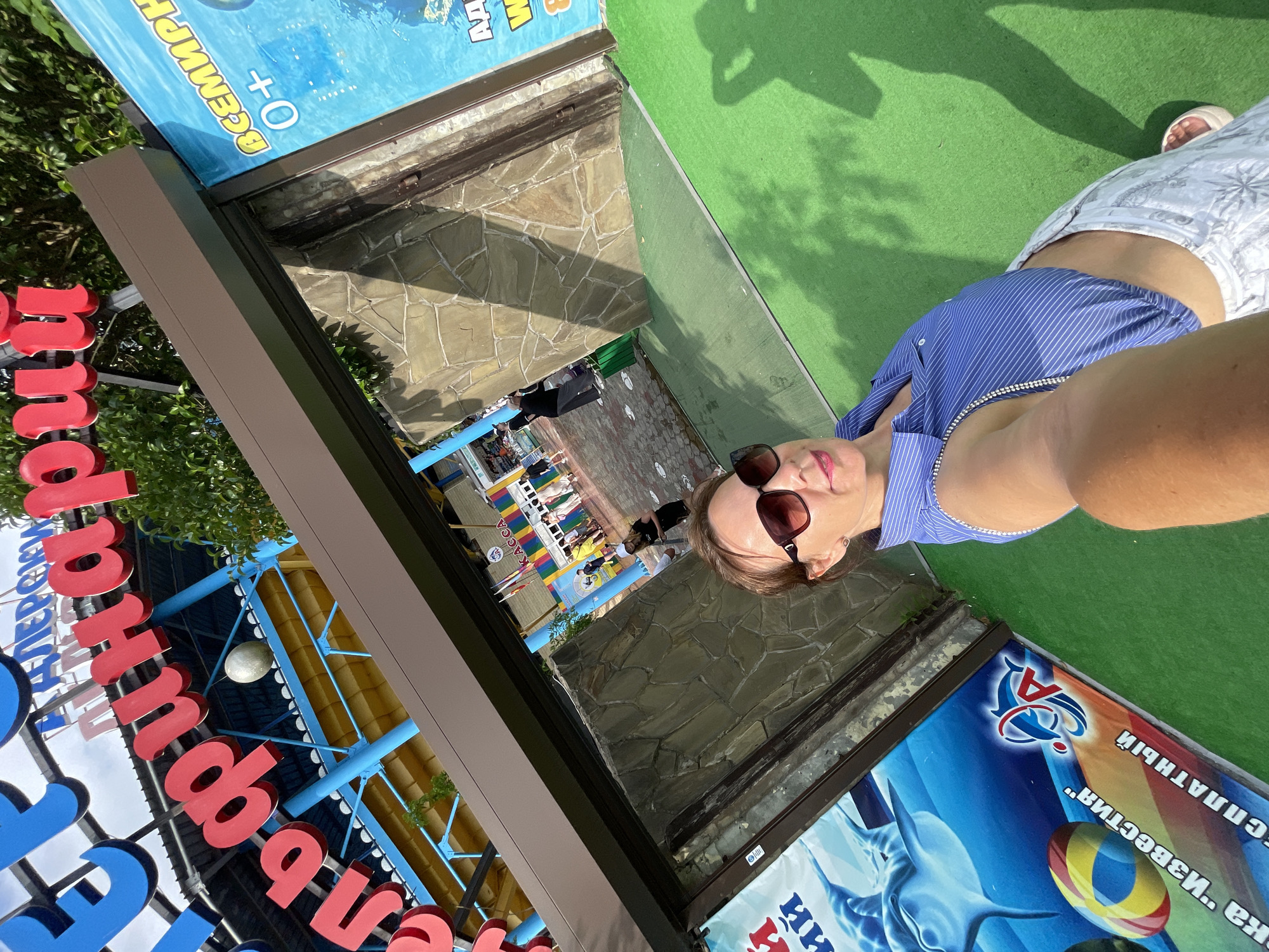 A woman is taking a selfie at the Sochi Dolphinarium. She is wearing sunglasses and a blue and white striped shirt. In the background, there is a large sign with Russian text that reads "Sochi Dolphinarium" and an advertisement for a water park. The ground is covered in green artificial turf. There are other people walking around in the background, as well as some trees and foliage. The sun is shining and casting shadows on the ground.