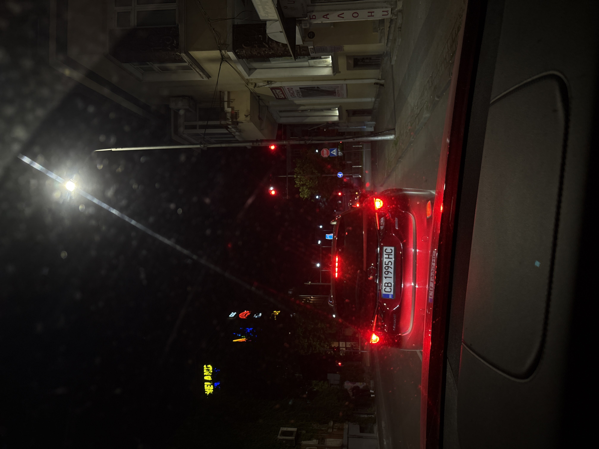 The image depicts a nighttime urban street scene, observed from inside a vehicle in Plovdiv, Bulgaria. The foreground shows the red dashboard and windshield of the viewer's car, with visible water droplets on the glass, suggesting wet conditions.
Directly ahead, a red car, likely a Mazda SUV, is stopped, displaying illuminated red taillights. Its Bulgarian license plate, "CB 1995 HC," is clearly visible. The vehicle is positioned at an intersection where traffic lights are red, indicating a stop.
To the right, a multi-story building with a light-colored facade, windows, and balconies is illuminated by spotlights. Several signs are visible on this building: one reads "SAVOY" above "ХРИСТО УРГИЙН" (Christo Urguin), and another vertically oriented sign on an adjacent storefront displays "БАЛОНИ" (Balloons) in Cyrillic. A tall street lamp illuminates the right side of the street and building.
On the left side of the street, darker buildings with visible air conditioning units and overgrown foliage are present. Two indistinct human figures are walking on the sidewalk near these buildings.
Further into the background, several vibrant neon signs are visible, including yellow-green text reading "НЕ! АНD" and a red sign with "CCC," along with other blurry colored lights. Street signs include a red octagonal "STOP" sign and a blue circular traffic sign. Pedestrian crossing markings are visible on the road surface.
