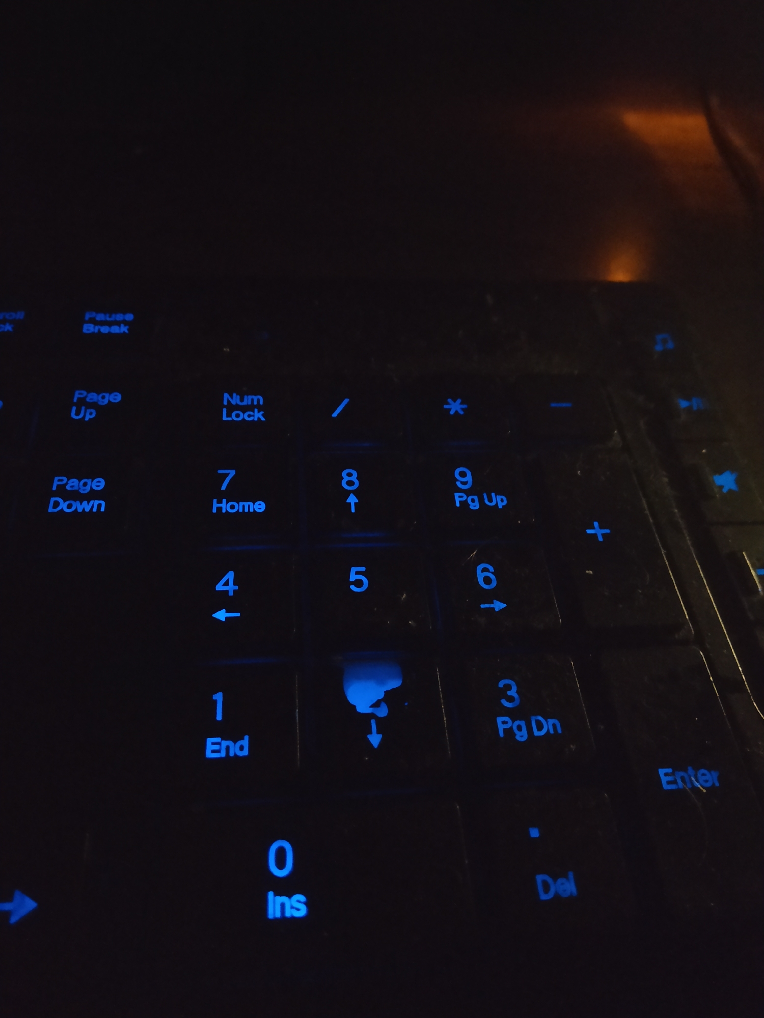 The image shows a close-up, low-angle shot of a black keyboard with illuminated blue keys. The keys are predominantly displaying numbers and function labels. Visible keys include "Pause Break," "Page Up," "Num Lock," "/", "*", "-", "Page Down," "7 Home," "8 ↑," "9 Pg Up," "4 ←," "5," "6 →," "1 End," "0 Ins," "Del," and "Enter." A faint orange glow from an unseen light source is visible in the upper right portion of the image. The keyboard appears to be resting on a dark surface, possibly a desk. There are no people or any indication of the city of Batumi, Georgia in the image. The scene is dark, with the primary illumination coming from the illuminated keys.
