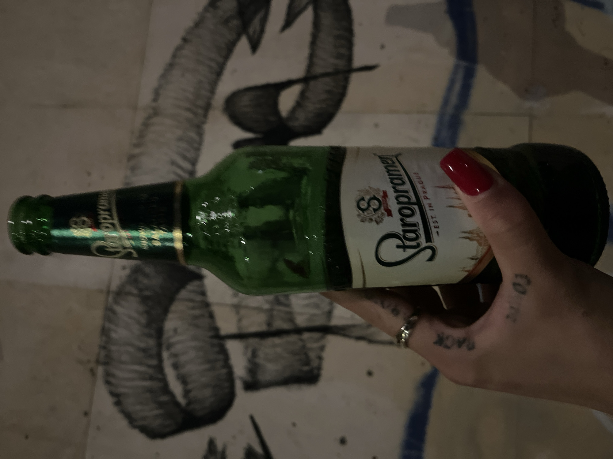 A hand with red fingernails and a silver ring holds a green bottle of Staropramen beer in front of a wall with graffiti. The graffiti appears to be a large black outline of lips. The hand has several tattoos, including the word "PACK" on the fingers and "21 03" on the palm. The background is out of focus, but appears to be a tiled wall.