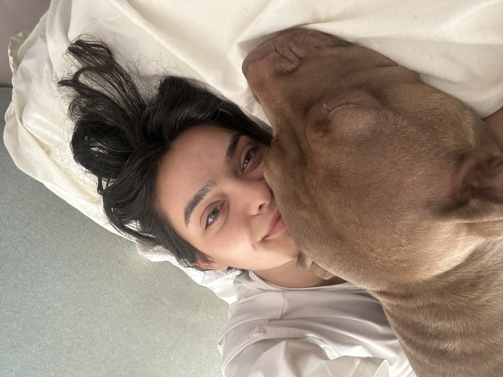 A woman and a brown dog are lying in a bed, which is made with white linens. The woman is on the right side of the image, with her head resting on a pillow. Her eyes are open, and she is looking at the camera. The dog is on the left side of the image, with its head resting on the woman's shoulder. The dog's eyes are closed, and it appears to be sleeping. The background of the image is a plain, light green wall. The overall impression of the image is one of intimacy and comfort.