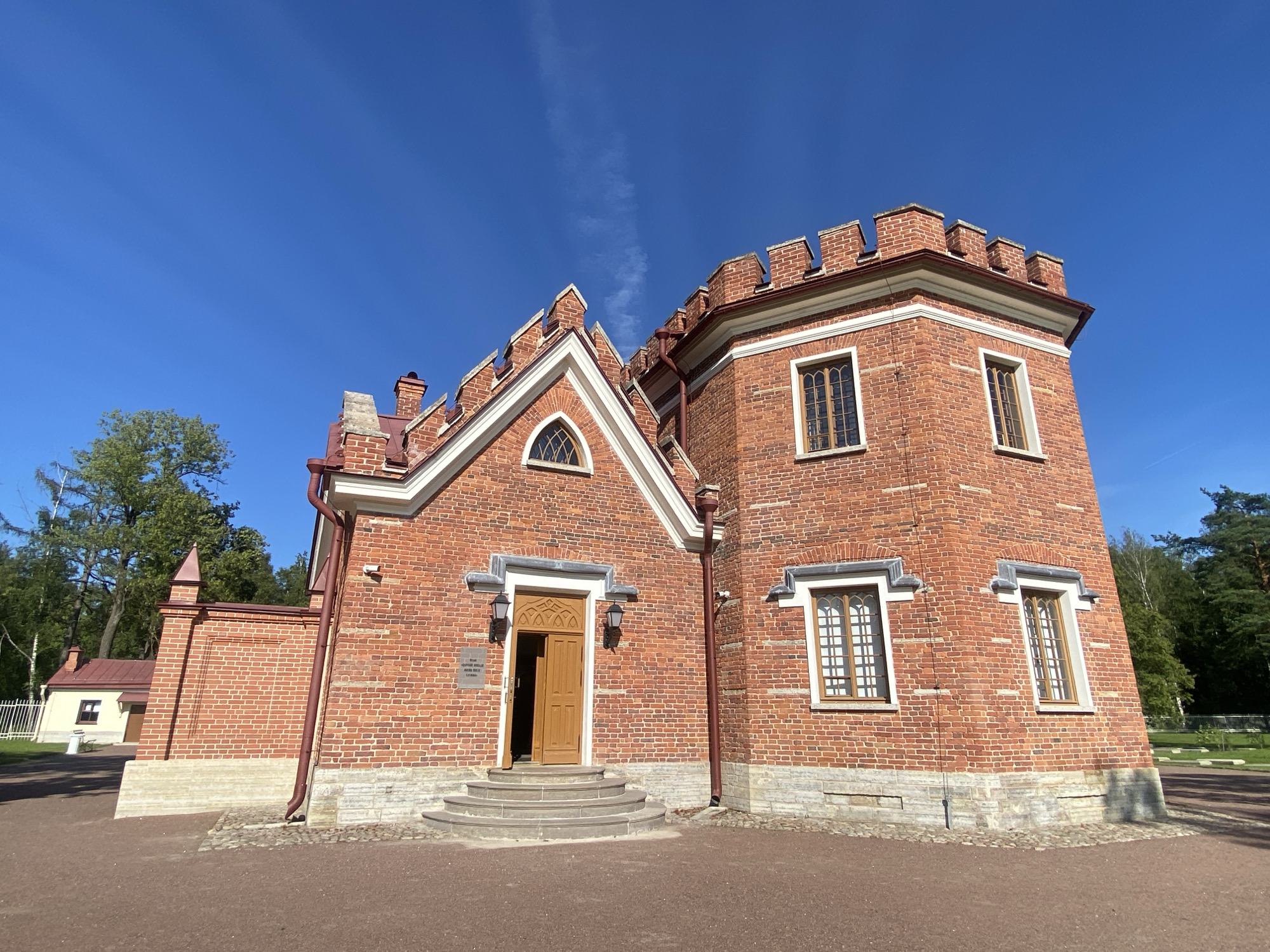 The image shows the Chinese Palace or the Small Chinese Palace in the Catherine Park in Pushkin, Russia. It is a bright, sunny day with a clear blue sky and a thin wispy cloud. The palace is a two-story brick building with a tower, designed in a picturesque style that imitates Chinese architecture. The red brick walls are decorated with white trim around the windows and battlements at the top of the walls, resembling a castle. The main entrance is a wooden double door with a semicircular arched opening, reached by a wide, shallow stone staircase with three steps. Two dark-colored lanterns flank the entrance. The windows are tall and narrow with intricate wooden lattice work. The surrounding grounds are covered with reddish-brown gravel, and there are some cobblestones near the base of the building. To the left of the palace, a low brick wall extends, with a small building with a red roof visible in the distance. Trees with green foliage are visible behind the palace and to the right, suggesting a park setting. The overall impression is one of a charming, historical architectural structure set within a well-maintained outdoor environment.