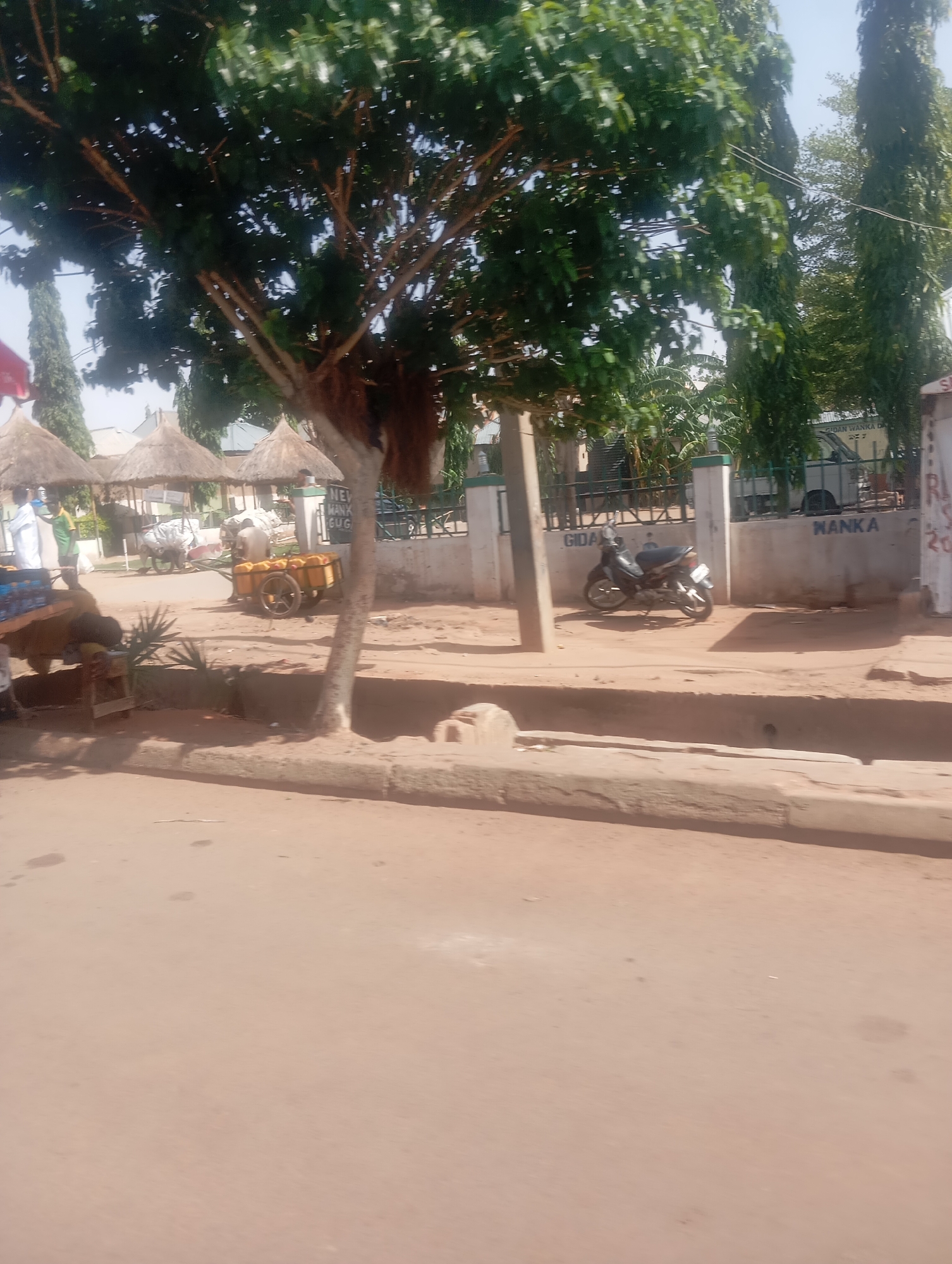 The image shows a street scene in Gombe, Nigeria. In the background, there are thatched-roof structures suggestive of market stalls or small businesses. A cart laden with yellow goods (possibly mangoes or other fruits) is visible, pulled by an animal. In the midground, a motorcycle is parked next to a low wall bearing the word "GIDA" and "MANKA". There is a tree in the foreground. The road surface is dirt or unpaved. In the background, the text "Gidan Wanka D" can be partially viewed. There is a building structure with green walls also in the background.