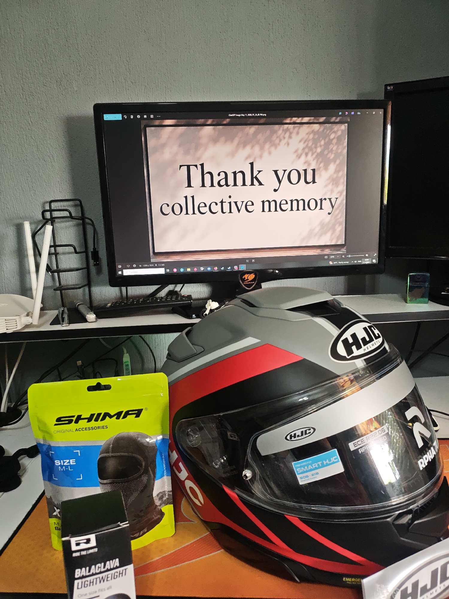 The image shows a desk with a computer monitor displaying a white rectangle with the text "Thank you collective memory" in a dark font. In front of the monitor, there's a gray, red, and black HJC motorcycle helmet. To the left of the helmet are packages from Shima and Ride the Limits containing balaclavas. A keyboard and a white Wi-Fi router are also visible on the desk, along with other accessories. The background wall is a pale gray color. The location is unknown.