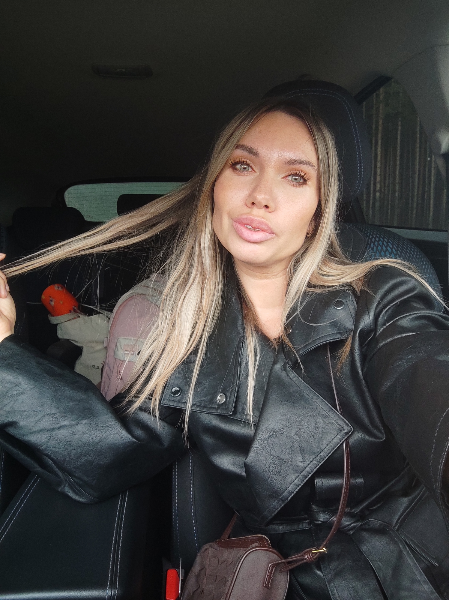 A woman is seated in the driver's seat of a car. She has long blonde hair and is wearing a black leather jacket. A small, patterned brown purse hangs from her left shoulder. A child's car seat is visible in the back seat, containing an orange and beige item. The location appears to be Votkinsk, Russia based on the visual cues.