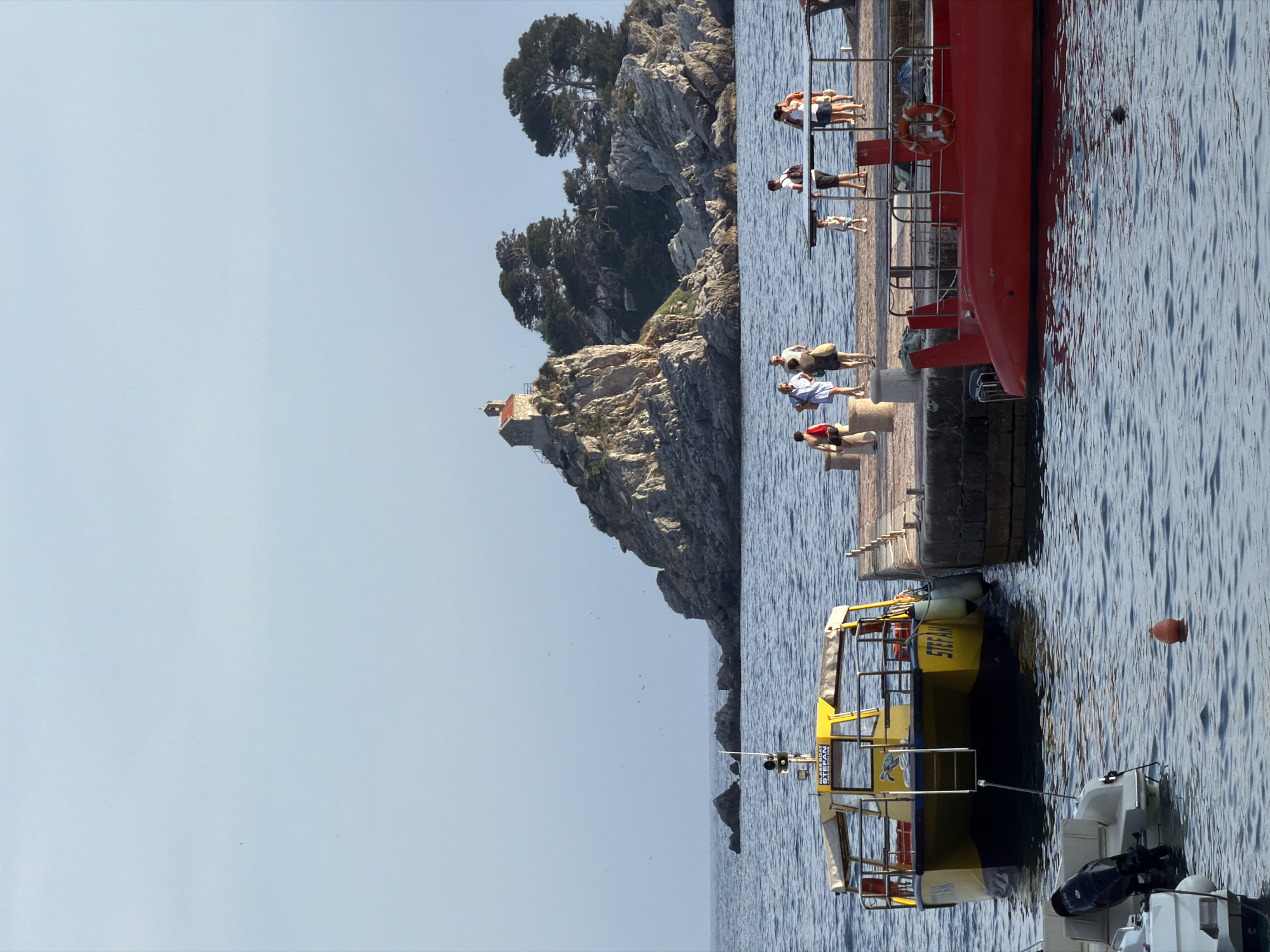 In Petrovac centar, Montenegro, the scene depicts a serene seascape under a clear, light blue sky. The calm, deep blue water is occupied by several vessels. In the foreground left, the stern of a white motorboat with a black outboard motor is visible. Adjacent to it, to the right, is a yellow catamaran-style boat named "STEFAN," featuring a covered seating area and a ladder leading into the water. Moored next to it is a bright red submersible, its sleek design contrasting with the yellow boat.
A concrete pier extends from the right side of the frame, where a group of people are gathered. Some are strolling, while others stand in conversation. A young child is seen walking on the pier. In the background, a rocky island rises from the sea, topped with dense green trees and a small, red-roofed building with a steeple, possibly a chapel. Several rocky outcrops are scattered in the water around the main island. The overall impression is a tranquil summer day at a coastal town.