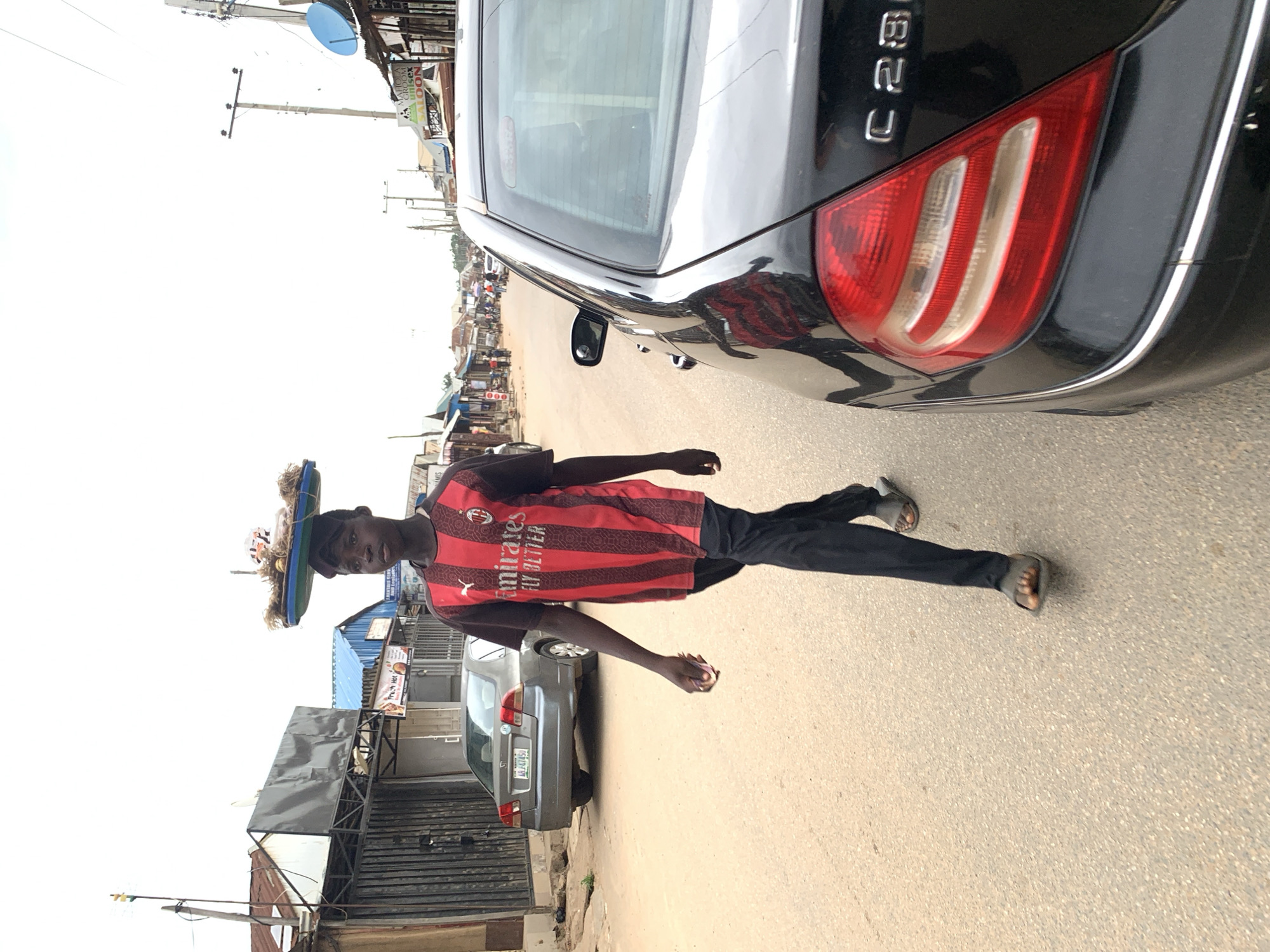 A young Black male, appearing to be in his late teens or early twenties, walks along a paved road in Abuja, Nigeria, carrying a tray of goods balanced on his head. He is wearing a red and black striped jersey with "Emirates Fly Better" written on it, along with black pants and dark sandals. The tray on his head contains what appears to be food items and straw. He is walking away from the camera, towards a row of roadside structures and a distant street scene.
To the left of the frame, a silver sedan is parked on the shoulder of the road. To the right, the rear of a black Mercedes-Benz C280 sedan is visible, parked on the same road. The environment appears to be a commercial or residential street with various buildings, some with corrugated metal roofs, lining the sides of the road. Power lines and a satellite dish are visible against a bright, overcast sky. The road surface is asphalt. The overall impression is a busy street scene in an urban or semi-urban area.