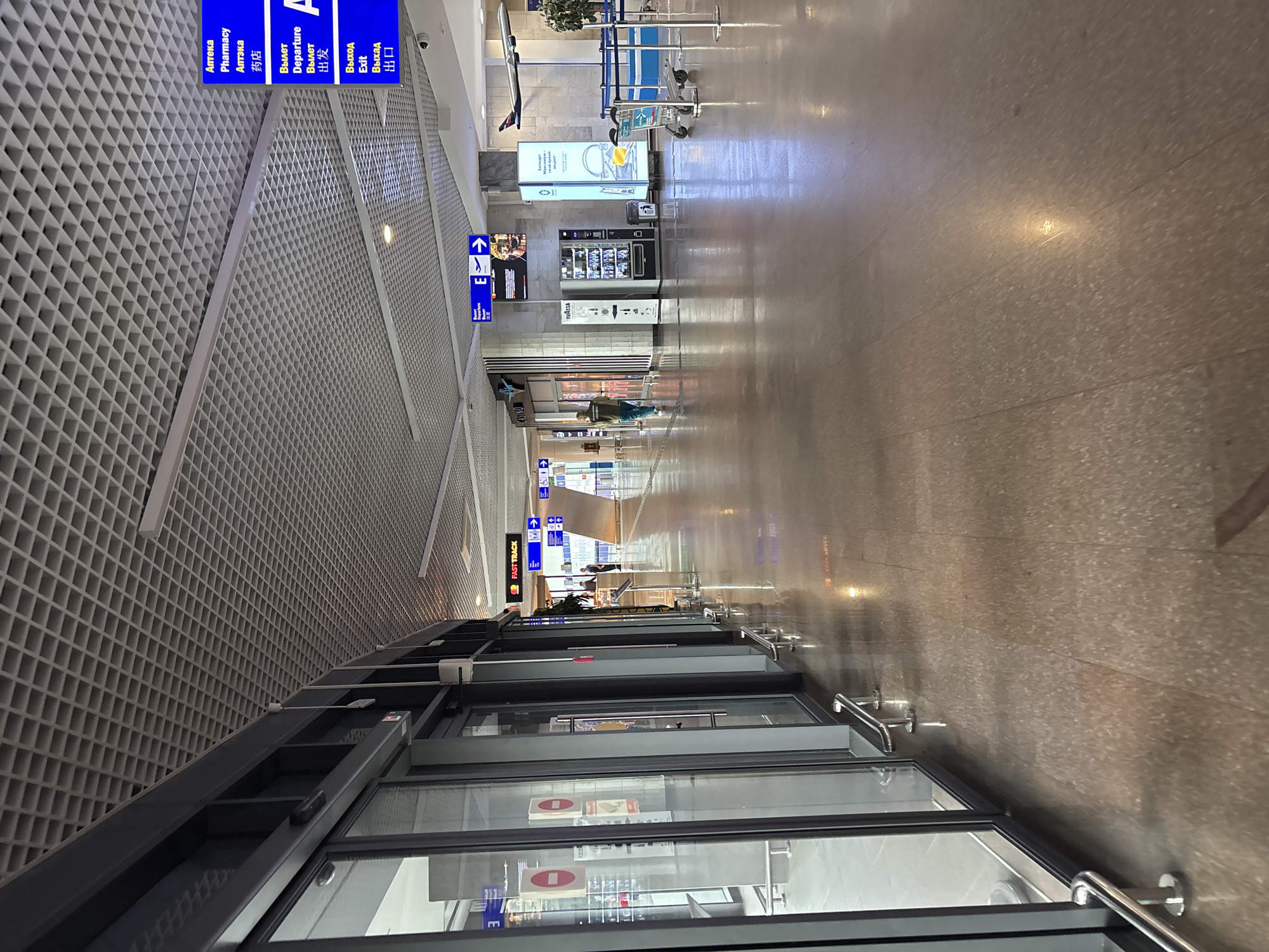 The image depicts an interior view of an airport terminal, likely in an area with shops and services. The perspective is from a hallway looking forward, with shops on the left and a wider corridor extending into the distance. The ceiling is a grid of acoustic tiles with embedded lighting.
On the left, there are large glass-fronted shop windows with dark frames, revealing displays of products and signage. Some of the signs appear to be red circles with a diagonal line, suggesting prohibitions.
In the center of the frame, the floor is polished and reflective, showing the overhead lights. A person wearing a dark jacket and light-colored pants is walking away from the camera down the corridor. Further ahead, on the right side of the corridor, there are various services and amenities. These include a vending machine, a sign with the word "CASINO", and a display advertisement featuring an airplane model. There are also signs in multiple languages, including Russian and Chinese, indicating "Pharmacy," "Departure," and "Exit." Blue directional signs with arrows are visible, pointing towards different areas.
Further down the corridor, there are queuing barriers with blue ropes, and luggage carts are present. A model airplane is suspended from the ceiling near the right wall. Plants in pots add a touch of greenery to the environment. The overall atmosphere suggests a functional and modern airport setting.
