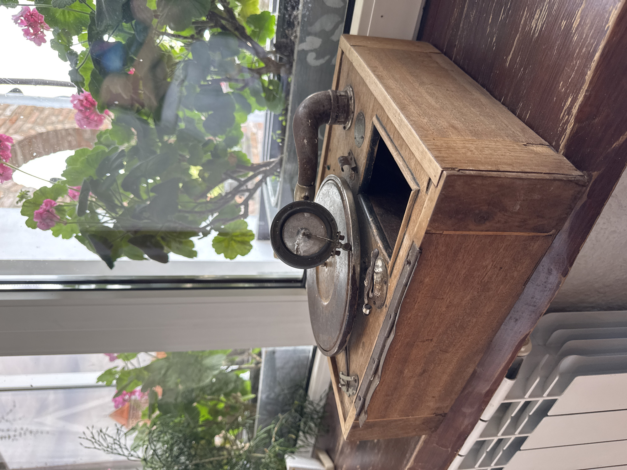 An antique gramophone sits on a wooden surface, positioned in front of a window with plants. The gramophone, made of wood, has a metal turntable and a horn with a needle. The window overlooks a garden with pink flowers and greenery. In the foreground, a white radiator is visible. The overall scene suggests a domestic setting, likely a home or a shop, in the city of Mirski Selski Savet, Belarus.