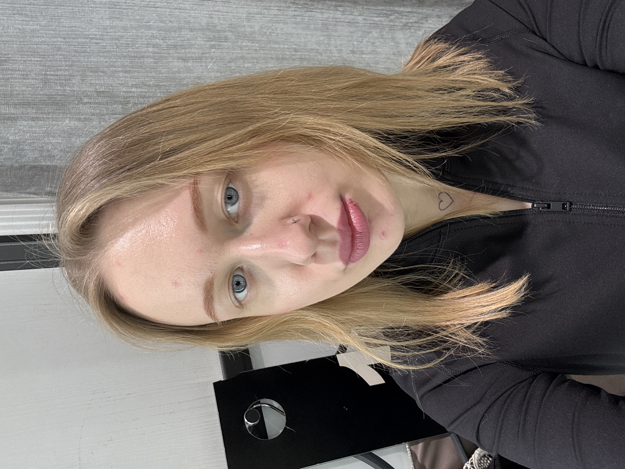 A woman with blonde hair and light blue eyes, wearing a black zip-up top, is facing the camera at a close angle. She has a small heart tattoo on her neck and a nose piercing. The background appears to be a wall with a light gray texture. Part of a black object is visible in the lower portion of the frame. The scene appears to be indoors. The location is not determinable from the image alone.