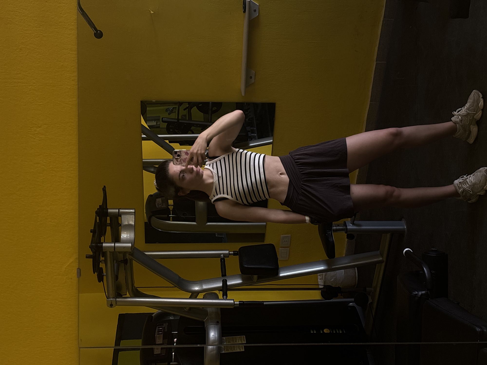 A young woman is positioned in front of a mirror in a gym setting in Unknown, Unknown. She is holding a mobile phone, apparently taking a self-portrait. The gym contains exercise machines with metal frames and black components. The room's walls are painted a yellow color.