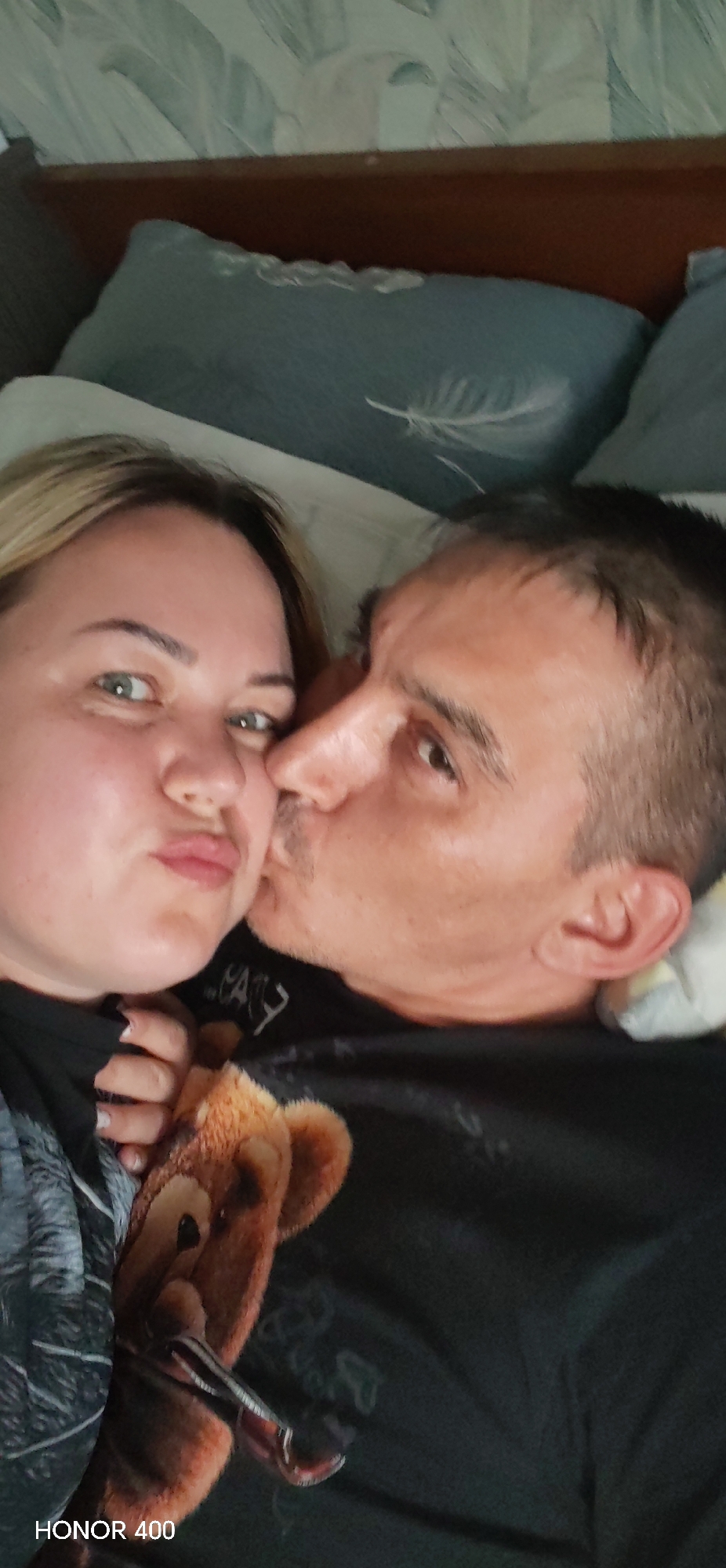 A selfie captures a woman with blonde hair and a man kissing her cheek. The woman, positioned on the left, has her lips pursed and holds a teddy bear. The man, slightly right, is kissing her. They are lying on a bed with light blue pillows. The photo was taken with an HONOR 400 phone. The scene appears to be indoors, likely in a private residence. There is no evident information related to the city of Noviy, Russia.