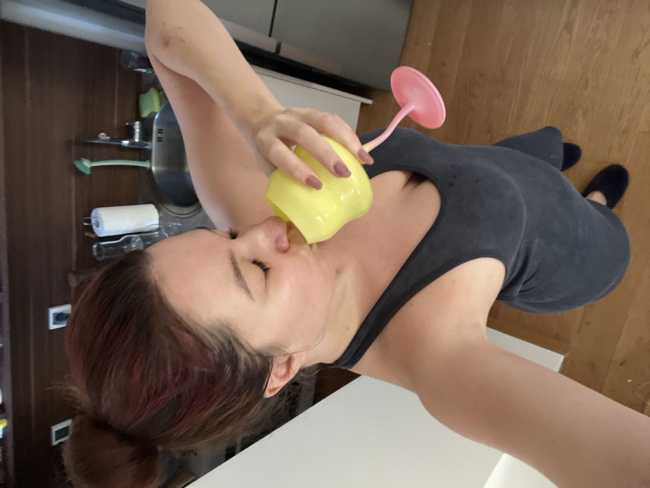 A female subject is depicted indoors within a residential kitchen or kitchenette setting. The image is captured from a high, downward angle. The subject, with reddish-brown hair pulled into a bun, is wearing a dark gray ribbed tank-top dress and dark slippers. She is actively drinking from a unique yellow glass with a pink stem and base, holding it with her right hand, her eyes closed. A red mark is visible on her upper right arm. The background includes dark wooden upper cabinets, a white electrical outlet, a stainless steel sink with a faucet, a green scrubbing brush, a green sponge, a roll of white paper towels, and a white refrigerator or tall cabinet. The floor is light brown wood. The exact location within Budva, Montenegro, cannot be determined from the visual information present in the image.
