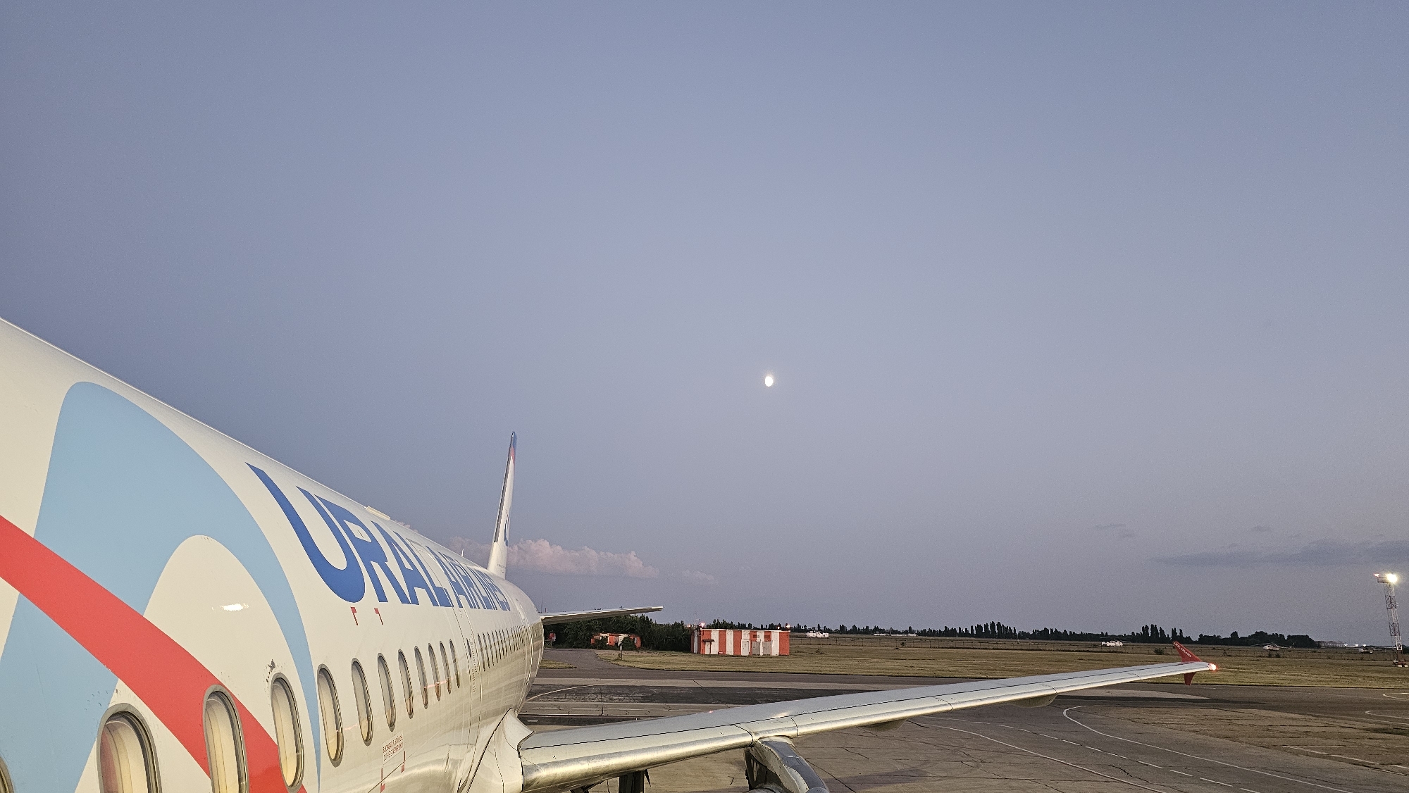 This is an outdoor, ground-level, wide shot of an airplane at an airport at dusk. The aircraft, an Ural Airlines plane, is parked on the tarmac, with its fuselage and wing taking up the left and bottom portions of the frame. The plane features a white body with blue and red livery. The sky is a gradient of light to dark blue with a small, bright moon visible near the center of the frame. In the background, there are some low buildings with red and white striped roofs, trees, and airport lighting towers. The tarmac is visible in the foreground and mid-ground, with a runway in the mid-ground. The scene appears to be at Krasnodar International Airport (Pashkovsky Airport) in Krasnodar, Russia, given the airline's branding. There are no people visible in the image.