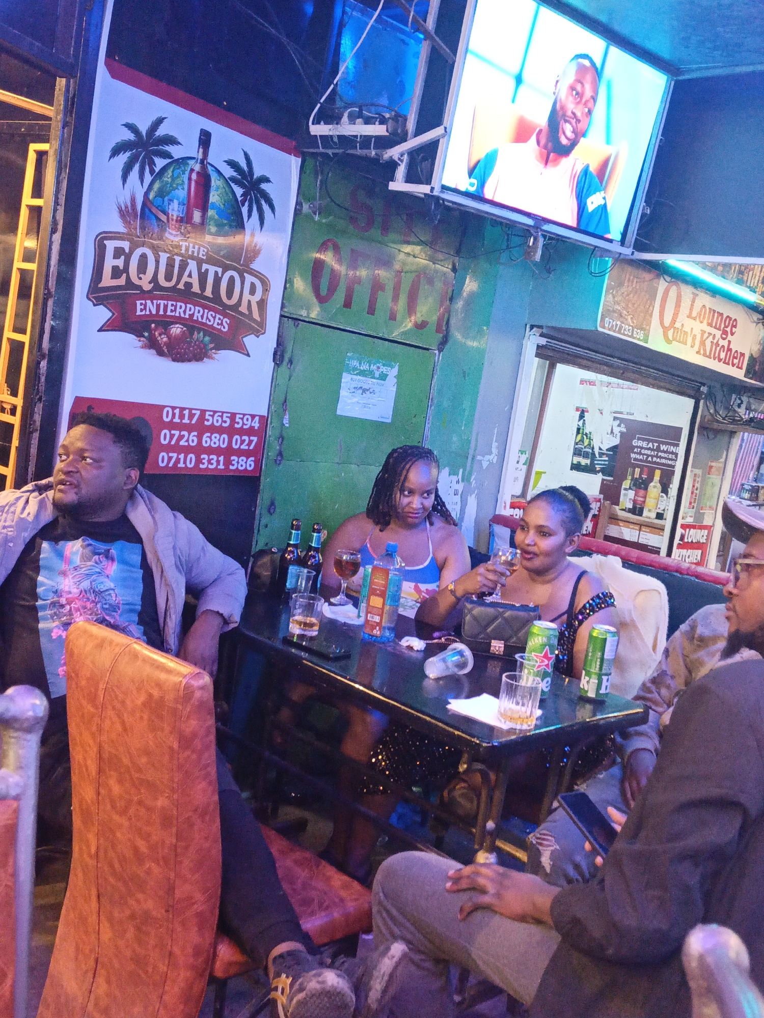 A group of four people are gathered around a black table in what appears to be a bar or lounge in Nairobi, Kenya. On the left side of the frame, a man with a dark t-shirt and a light-colored jacket is seated in an orange chair. Across from him are two women: one in a blue tank top and another in a black, sequined top, both holding glasses and engaged in conversation. The fourth person, on the right, is a man in a black jacket looking at a cell phone. The table is cluttered with beer bottles, cans, glasses, and other items. Overhead, a TV screen displays a man's face. Signage for "The Equator Enterprises," "Q Lounge Culn's Kitchen," and telephone numbers is visible in the background, suggesting the location. The environment appears to be a typical urban establishment.