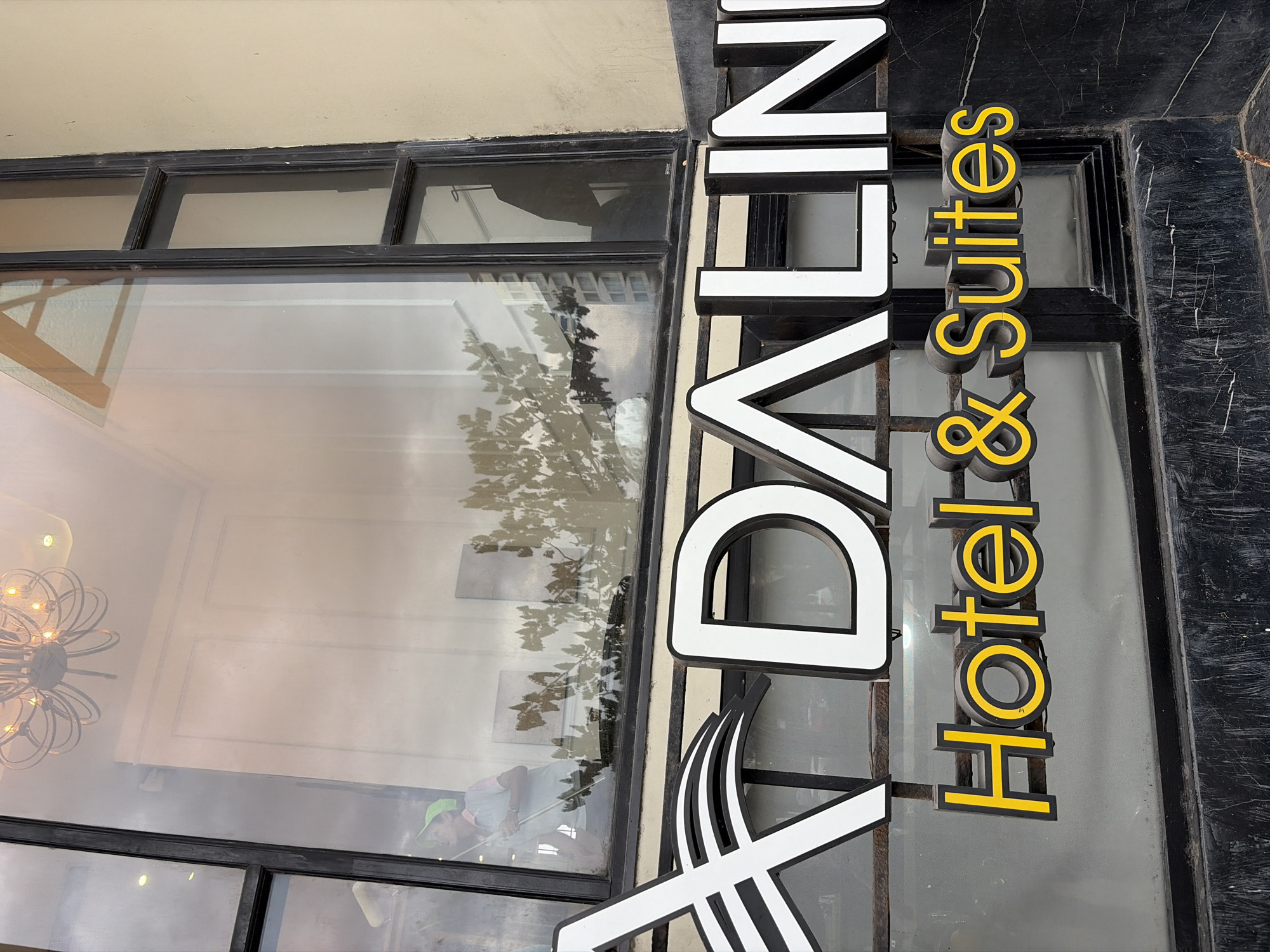 The image shows the facade of a hotel named "DALINI Hotel & Suites" in Da Nang, Vietnam. The hotel's name is displayed prominently on a large, illuminated sign. The sign features white, three-dimensional letters with black outlines for "DALINI" and yellow letters with black outlines for "Hotel & Suites". The hotel appears to be located in a commercial area, as suggested by the large window, the building's architecture, and the reflection of trees and buildings in the window. Inside the window, a person wearing a green cap is visible, possibly cleaning or working. The building's exterior is light beige, contrasting with the dark window frames and the dark, textured material below the sign. The lighting in the image suggests it was taken during the daytime, possibly with the sun reflecting off the building.