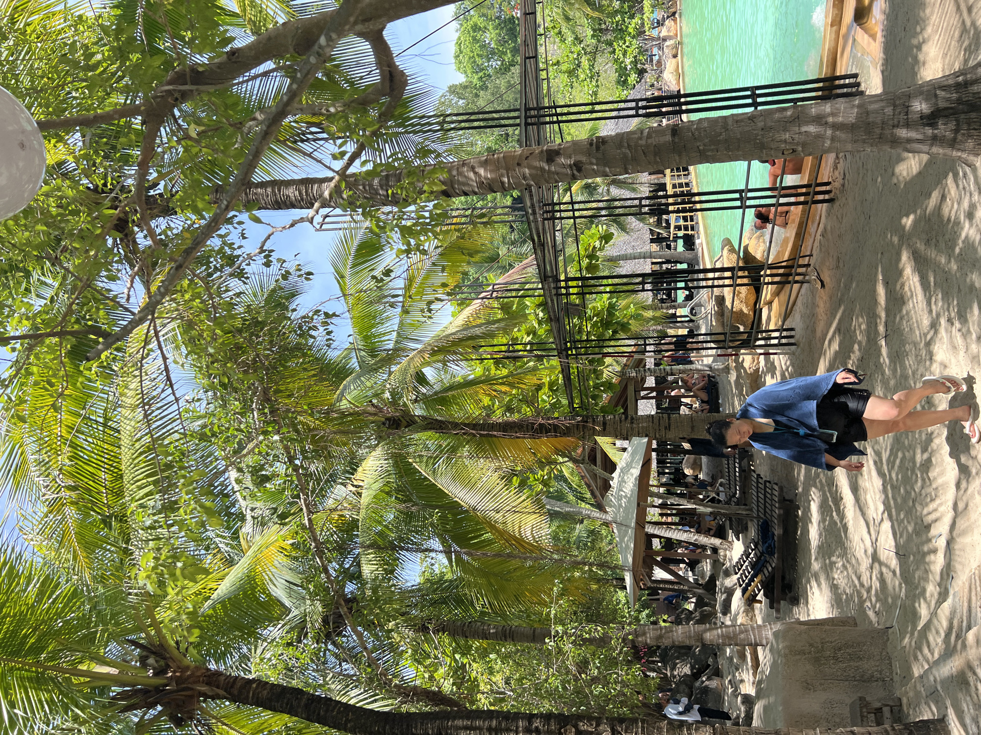 The image depicts a tropical resort setting in Tay Nha Trang Ward, Vietnam. A woman wearing a blue towel draped over her shoulders and black shorts walks across a sandy area. In the background, a turquoise swimming pool is visible, with several people relaxing by the poolside and in the water. Palm trees and lush greenery surround the area, casting dappled shadows on the sand. Several lounge chairs are arranged under thatched-roof structures. The scene suggests a relaxed atmosphere at a vacation destination.