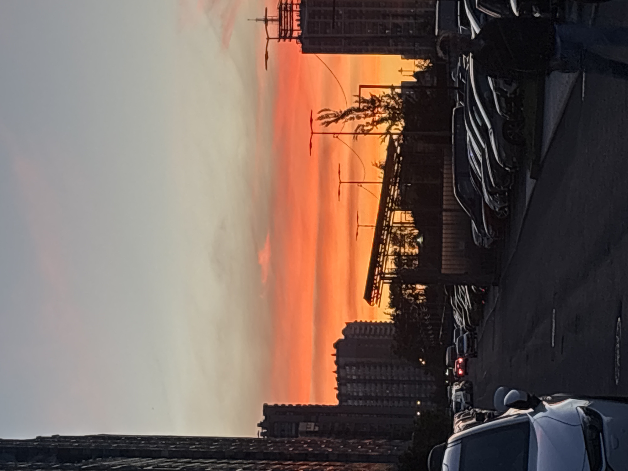 A vertical, medium shot captures a city street at dusk. The sky is ablaze with a vibrant orange and pink sunset, transitioning to a pale blue at the top. In the foreground, the front of a white car is visible on the left. The street is lined with parked cars, extending into the background. Several multi-story buildings dominate the cityscape on either side of the street, their dark silhouettes contrasting with the bright sky. On the right side of the image, a person wearing glasses and a dark jacket stands in the middle ground, facing away from the camera. Lampposts are scattered throughout the scene, some illuminated. A construction site with visible scaffolding is also present. The overall impression is of an urban environment at the end of the day.