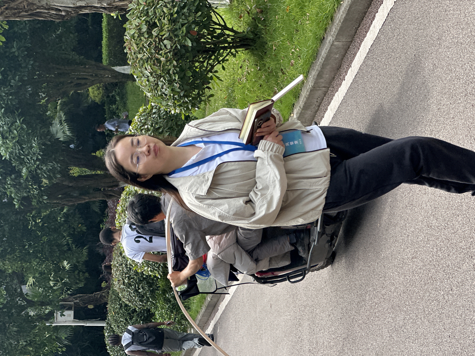 The image shows a young woman in Jiangjin, China, sitting on a vehicle on a paved surface, possibly a road or pathway. She wears glasses, a light jacket over a white shirt with a blue lanyard, and dark pants. She holds a book in one hand and a small blue object in the other. Several other people are present in the background, including one wearing a baseball jersey with the number 2 on the back. Lush green vegetation lines the side of the paved area, including trees and bushes.