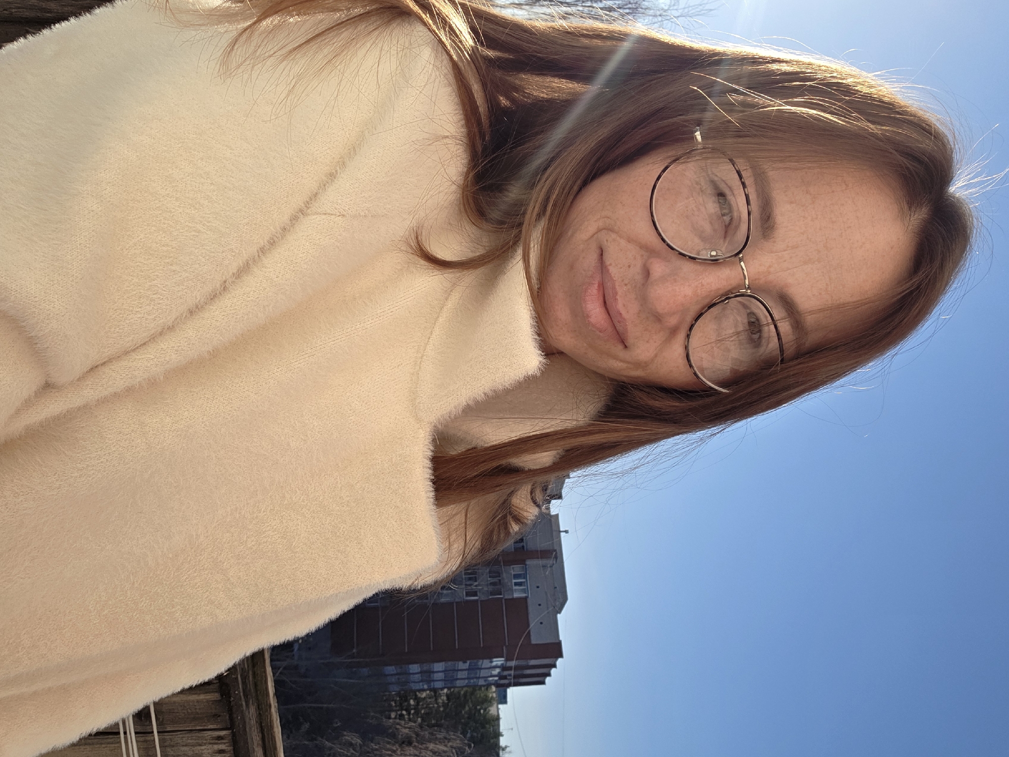 The image is a selfie taken by a woman with light brown hair and glasses. She is wearing a fluffy, light-colored coat. The background features a clear blue sky and a multi-story brick building, likely an apartment complex. The location is Chaikovsky, Russia, based on the building style common in Russian urban areas. The woman is positioned outdoors, possibly on a balcony or near a fence.