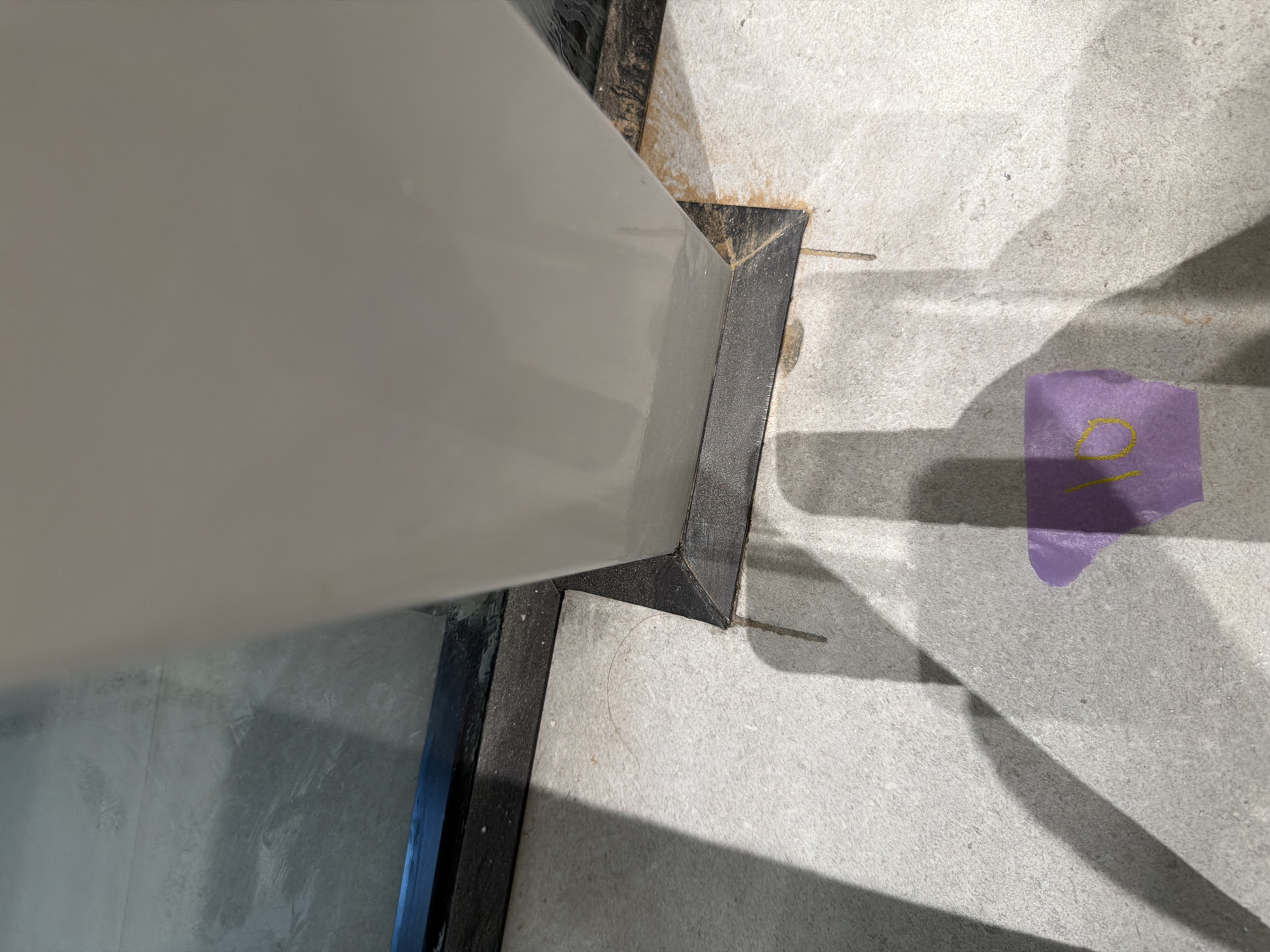 A close-up, low-angle shot shows the base of a large, off-white, rectangular column, its bottom edge fitted with a dark, angled metal trim that meets the light-colored tiled floor. The trim is secured to the column by a black border. On the floor, near the base of the column, a piece of purple tape with a yellow number "10" written on it is visible. Shadows of the column and what appears to be a window or glass partition are cast across the tiled floor, creating strong diagonal lines. The floor tiles have a speckled, light gray appearance. In the upper left, a reflection of what appears to be a large window with a dark frame is present. The scene appears to be indoors, possibly in a modern building or public space in Jerusalem, Israel. The lighting suggests an overhead light source.