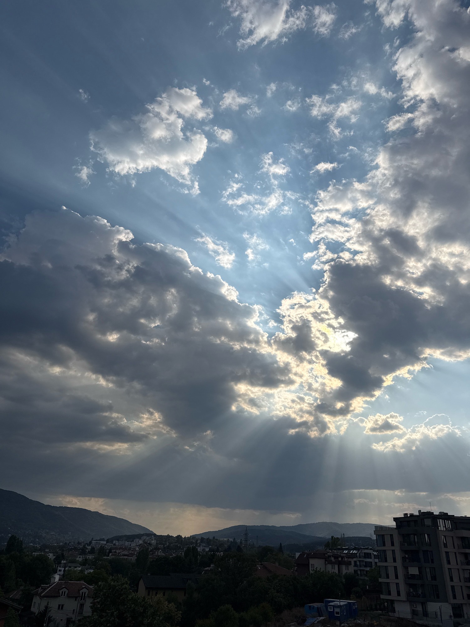 The image captures a dramatic sky filled with crepuscular rays emanating from behind scattered clouds. The sun's rays pierce through gaps in the clouds, creating luminous beams that illuminate parts of the sky and cast shadows. The clouds are a mix of dark, heavy masses and lighter, more wispy formations. Below the sky, a cityscape is visible, characterized by rolling hills in the background. The foreground features buildings of various sizes, including what appears to be a modern, multi-story apartment building on the right side of the frame. Trees and residential houses are interspersed throughout the urban landscape. The overall impression is one of a dynamic sky over a city during what appears to be either sunrise or sunset, with the light creating a striking contrast between the illuminated and shadowed areas. The location is identified as Sofia, Bulgaria, suggesting the urban setting is within this city.