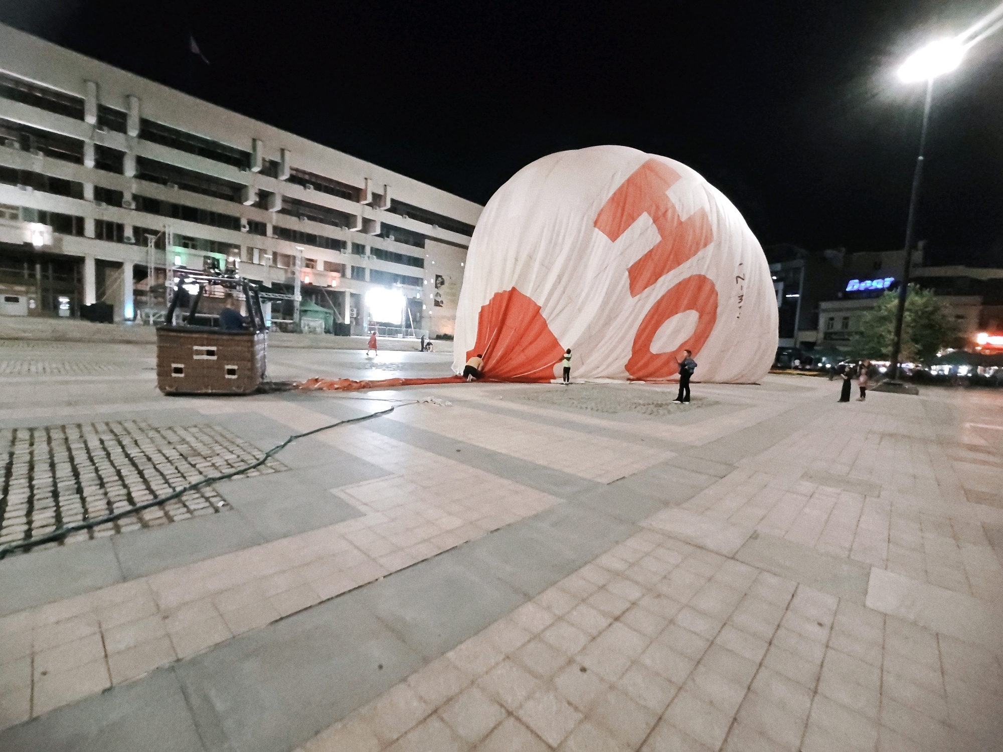 In an urban plaza at night, a large, partially deflated hot air balloon lies on the ground. The balloon is predominantly white with large, red lettering that spells "ON". Several people are visible near the balloon, some appearing to be working with it or observing. In the foreground, a wicker hot air balloon basket is positioned on the paved ground, connected to the balloon by a line. The plaza is paved with square tiles. In the background, a multi-story building with numerous windows and air conditioning units is illuminated. Streetlights and other building lights are visible, indicating a nighttime setting. The overall scene depicts a preparation or deconstruction phase for a hot air balloon event in a public urban space.
