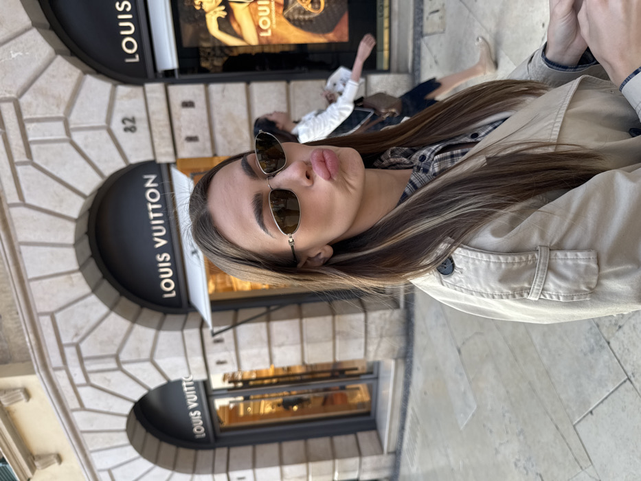 A female subject is positioned in the foreground, facing the camera with a "duck face" expression, lips pursed, and wearing dark, oval-shaped sunglasses with thin metallic frames. She has long brown hair with blonde highlights and is dressed in a beige trench coat over a plaid shirt.
The scene is set on a paved pedestrian sidewalk in front of the Louis Vuitton store located at Via Giuseppe Mazzini, 82, Verona, Italy. The store's classical stone facade features two prominent arched entrances/display windows. Above each arch, a black sign displays "LOUIS VUITTON" in white capital letters. The building number "82" is visible between the two arches. The interior of the store is brightly illuminated, showcasing merchandise.
In the mid-ground, partially obscured by the foreground subject, a second female is seen walking from right to left. She wears a white top, a dark lower garment, and light-colored shoes, carrying a small, light brown handbag on her right shoulder. The setting is an urban street under natural daylight.