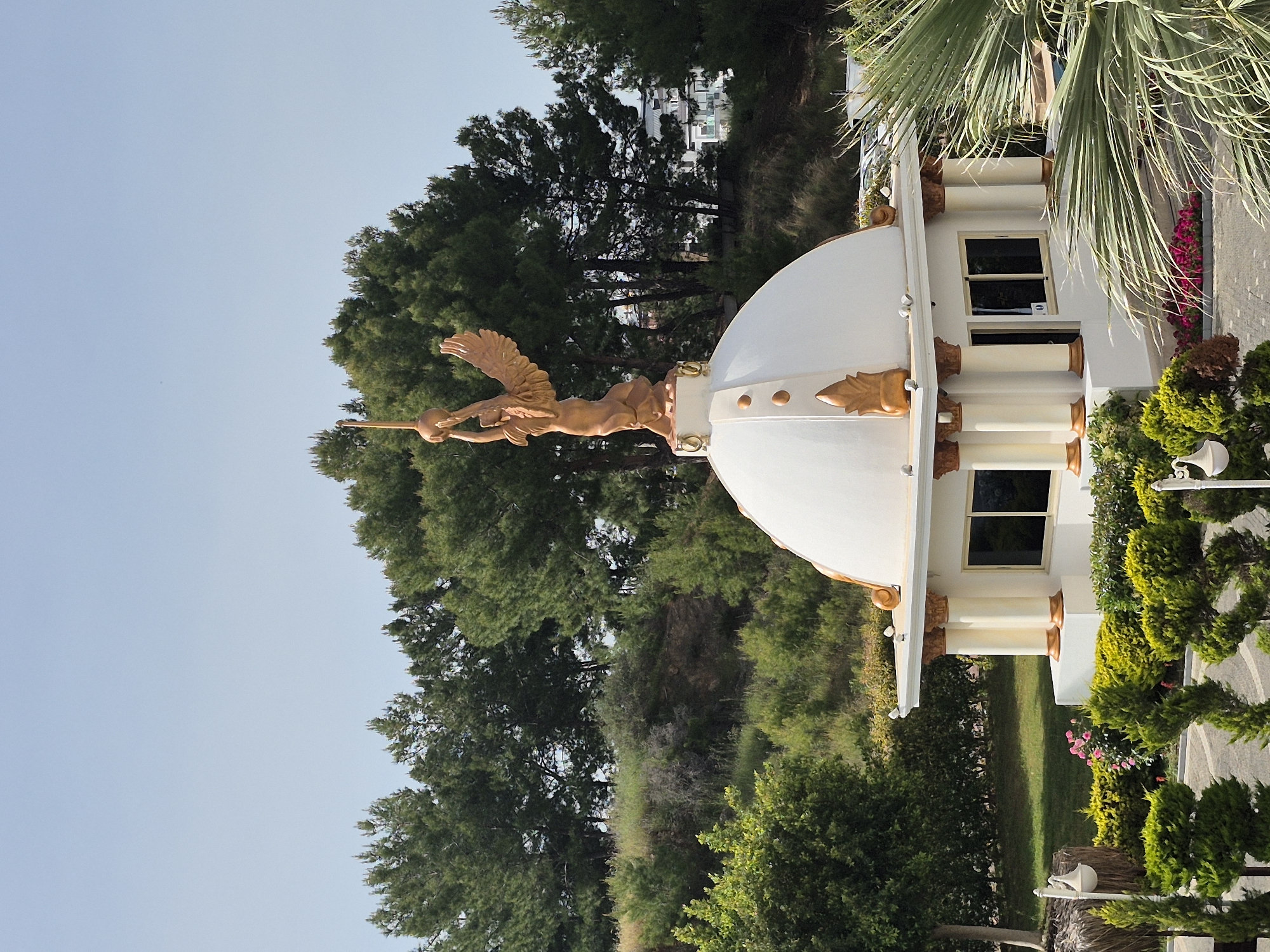 The image depicts the Kültür Merkezi in Alanya, Turkey. The white, domed building features architectural details highlighted in gold. Atop the dome sits a golden statue of a winged figure holding an object aloft. The building is surrounded by lush greenery, including trees, shrubs, and flowers. A palm tree is visible on the right side of the frame. The sky is clear and blue.