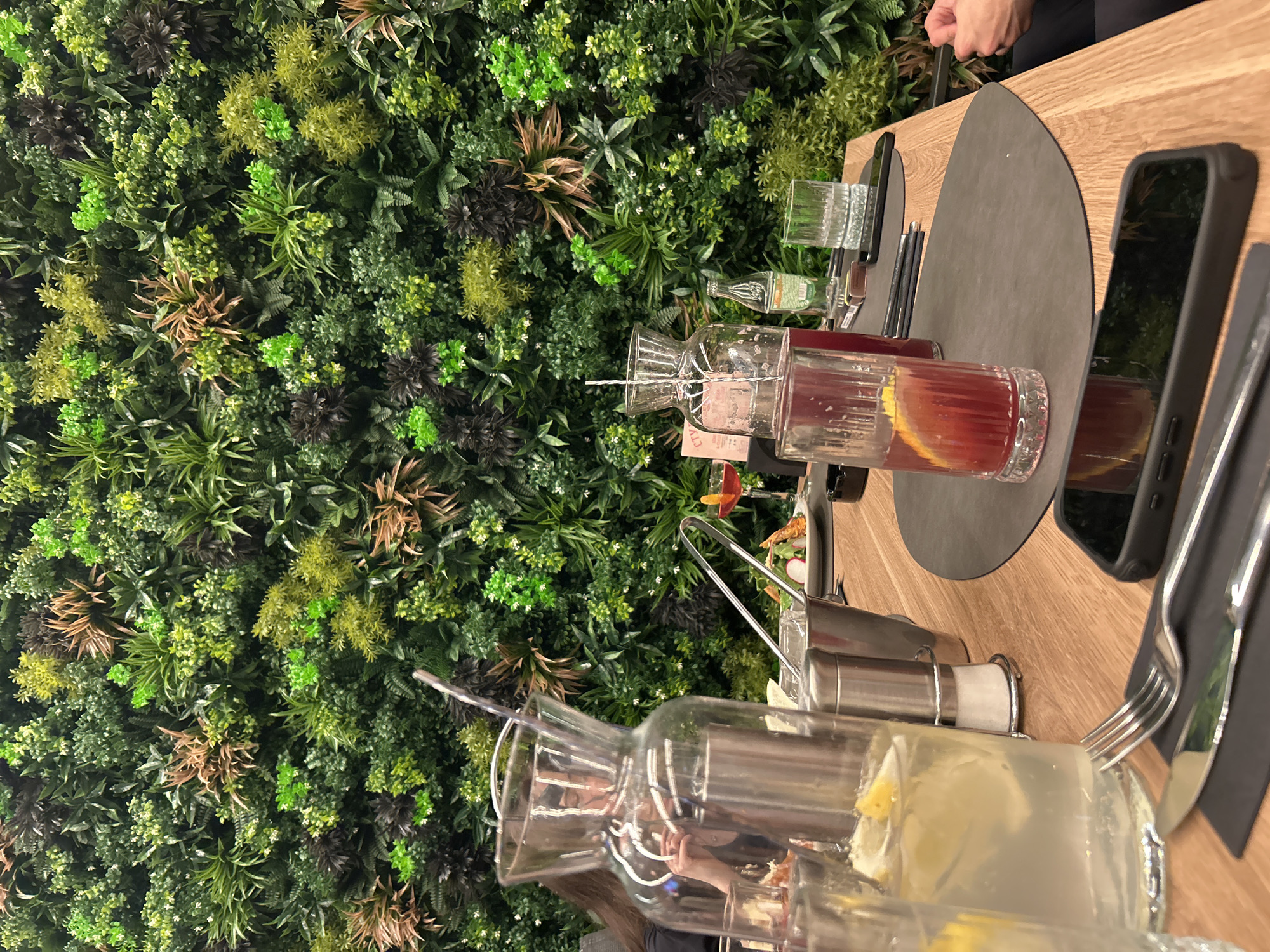 The image captures a table setting indoors, likely in a restaurant or cafe. A lush green wall serves as a backdrop, featuring various plants and greenery. On the table, there are several pitchers of beverages; one appears to contain a red fruit drink with citrus slices, and another contains a lighter beverage, possibly lemonade, with lemon slices. Glassware is visible, suggesting a drink service is available. Cutlery, including a fork and knife, is placed on a dark-colored placemat, along with a smartphone. The table itself is a light-toned wood. The scene is indoors, possibly a dining area with a natural aesthetic. The location is not discernable from the image alone.