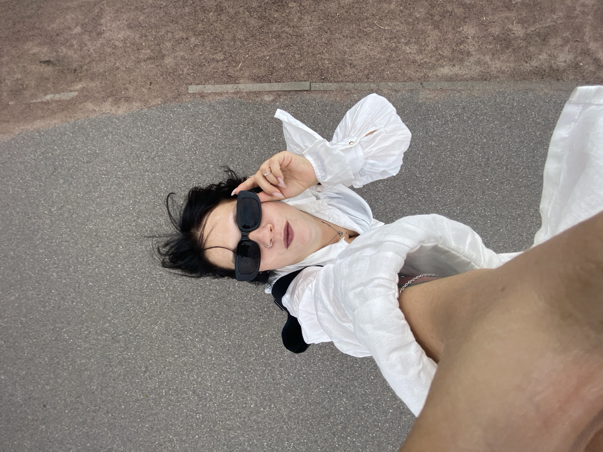 A person with dark hair and sunglasses stands on an asphalt surface. They are wearing a white collared shirt with puffed sleeves and a dark vest over it. The person is holding their hand up to their sunglasses. The asphalt surface is adjacent to a reddish-brown border. The perspective is from above, looking down. The scene appears to be outdoors, possibly in a city setting. The specific location in Saint Petersburg, Russia, cannot be determined from the provided image.