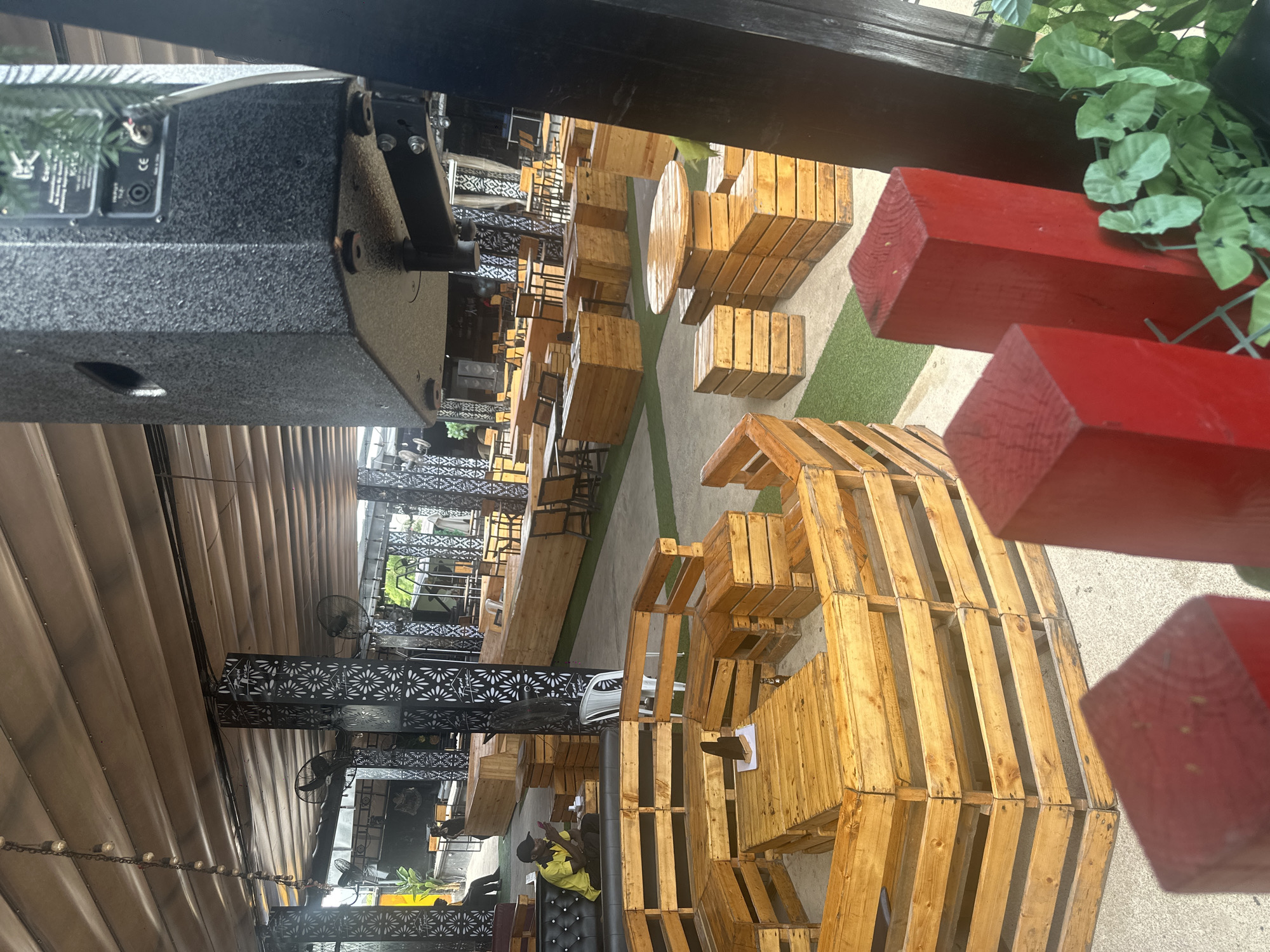 The image shows an outdoor seating area at a venue, likely a restaurant or bar, in Howayi, Nigeria. The seating is primarily composed of light-colored wooden crates and pallets, arranged to form benches and tables. In the foreground, there are red wooden posts. The background reveals more seating areas with similar wooden furniture, as well as tables and chairs. A large black speaker system is prominently visible hanging from the ceiling. The ceiling itself is covered with a canvas-like material, supported by pillars with intricate black metalwork. A person wearing a yellow shirt and a black cap is seated on a black sofa, looking at their phone. The overall environment suggests a casual, open-air gathering space.