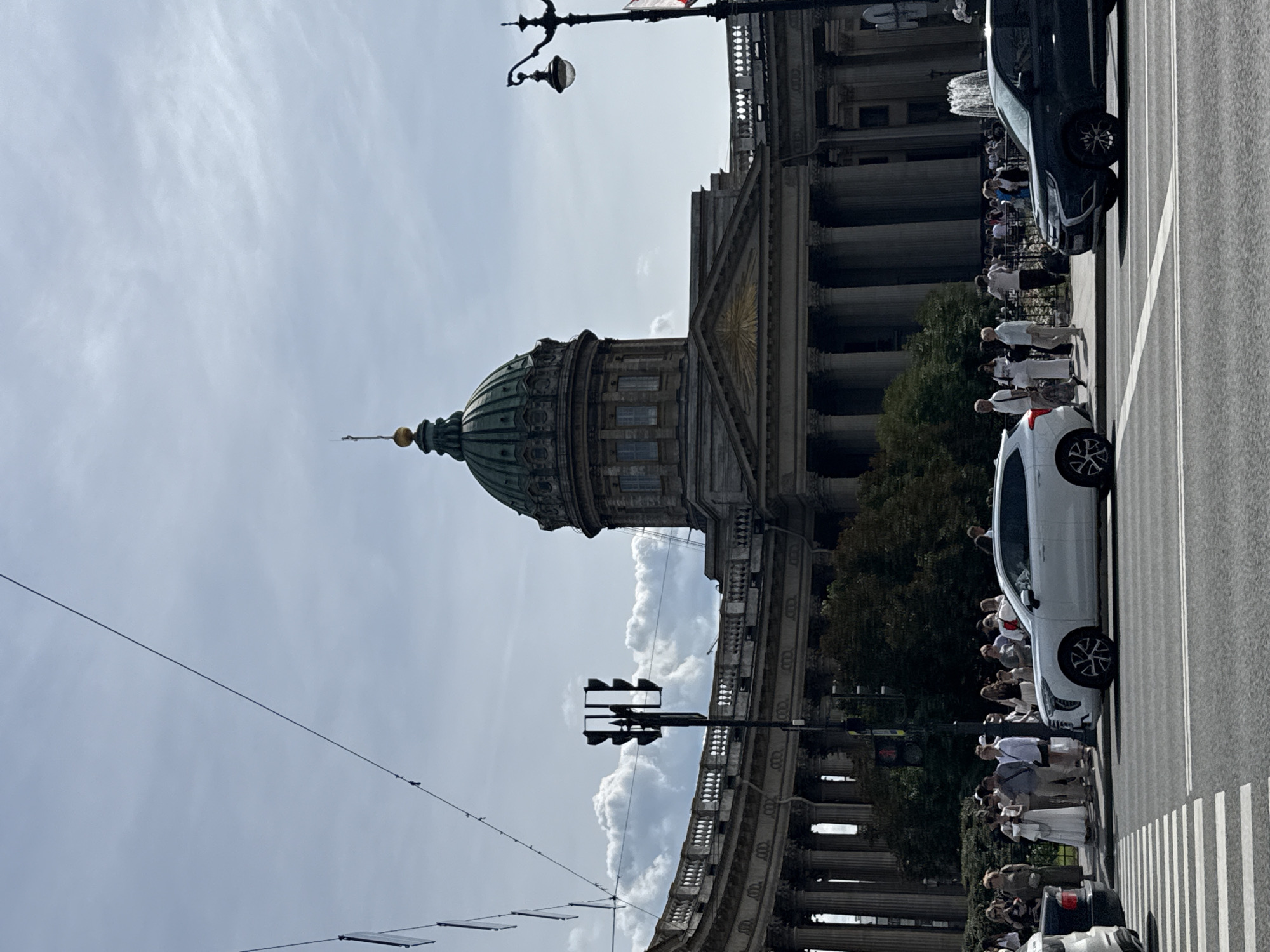 This is a daytime, medium-shot photograph of Kazan Cathedral in Saint Petersburg, Russia. The grand neoclassical building features a prominent green dome, colonnades, and detailed architectural ornamentation. In the foreground, a crosswalk and traffic lights are visible. Several cars, including a white SUV and a dark SUV, are on the road. A crowd of people, dressed in casual attire, are standing on the sidewalk, some waiting to cross the street. Trees and a fountain are also present in the scene, adding to the urban environment. The sky is partly cloudy, with dramatic cloud formations adding depth to the background.