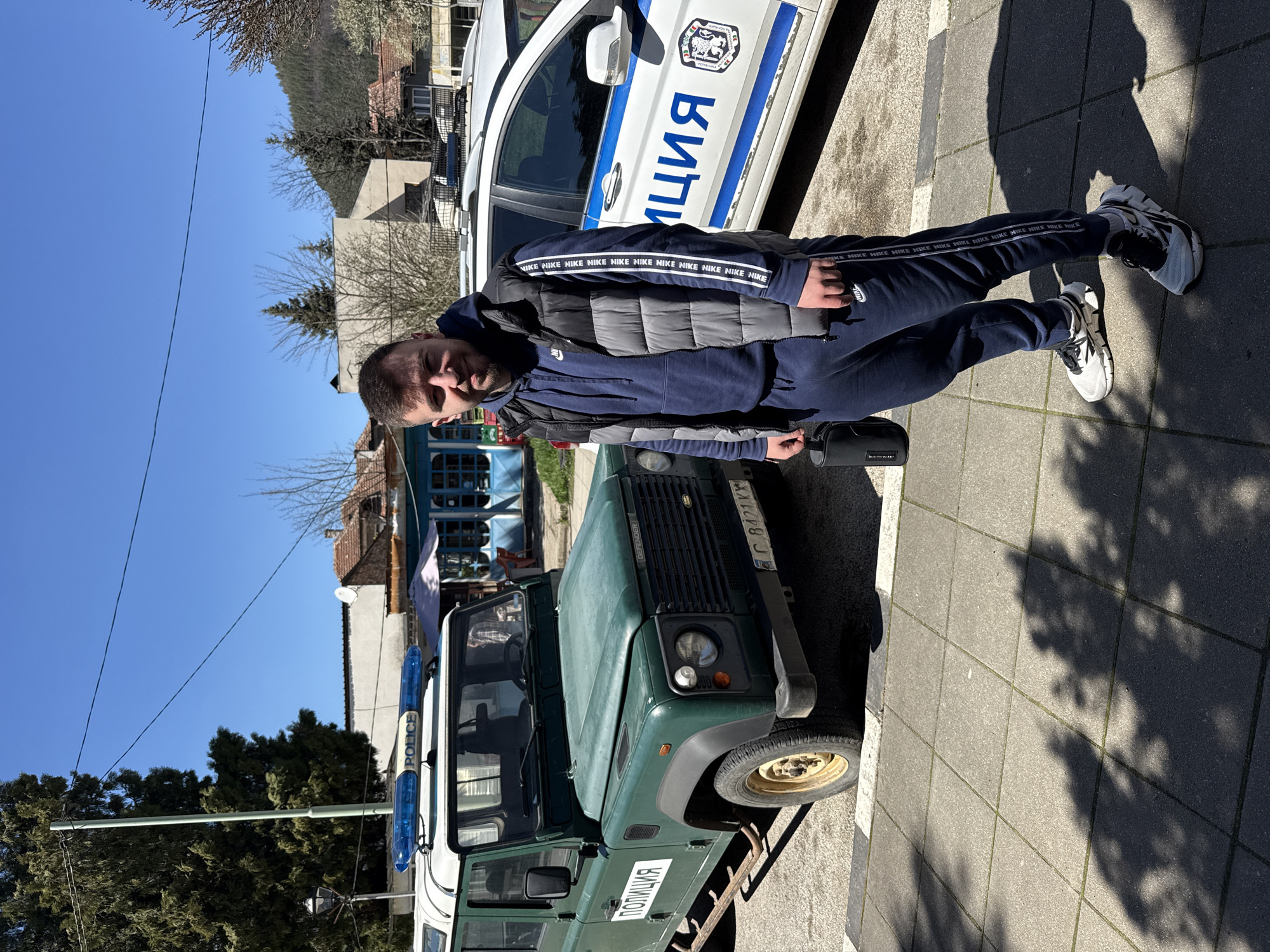 The image shows a man standing next to a green Land Rover Defender police vehicle and a white police car in what appears to be Etropole, Bulgaria. The man is wearing a dark blue tracksuit with white Nike branding on the sleeves and legs, a gray puffer vest, and athletic shoes. The Land Rover Defender has "ПОЛИЦИЯ" (Police) written on the side in Cyrillic and a Bulgarian license plate "C 8421 KX." The white police car also has the word "ПОЛИЦИЯ" displayed in Cyrillic. The setting appears to be an urban area with buildings and trees in the background. The sky is clear and blue, suggesting it is daytime. The vehicles are parked on a paved surface.