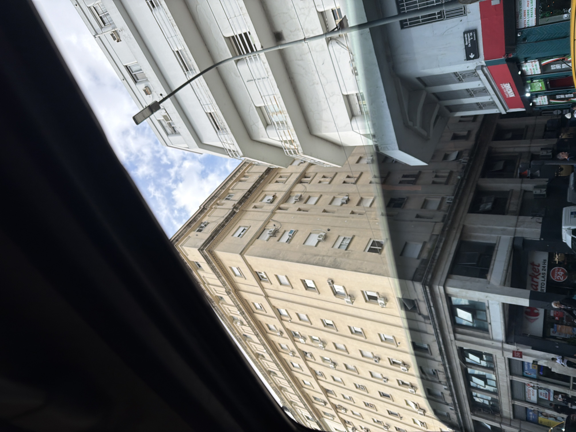 The image shows an urban scene, likely taken from inside a vehicle. The buildings are reflected in the glass window. There are two buildings in view. One is white, with what appears to be residential balconies. Adjacent to this building is a building with beige siding and multiple windows. Air conditioning units are visible on the facade of the building. The windows are mirrored on the opposite building, indicating the view is from a reflection.
On the right of the picture is a building with several store fronts, including one that reads “market” and “RTO LAS 24 Hs” (open 24 hours). The second store is “MANGIARE” with “PASTA LIBRE”, “PASTA LIBRE” appearing in stacked layers.
There are people on the sidewalk to the right of the photo.
The sky is partly cloudy and visible on the left of the photo. A street light is visible on the left as well.