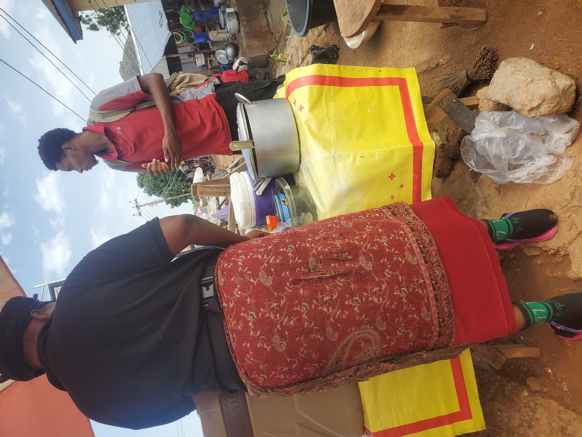 In Gwong, Nigeria, a person wearing a black shirt and a patterned red skirt stands at a street food stall. A young man in a red and gray polo shirt, carrying a backpack, stands to the right of the stall, looking away from the camera. The stall is covered with a yellow tarp and displays various cooking pots and containers. In the background, other market stalls and people are visible, suggesting a busy marketplace environment. The ground is dirt, and there is a broom and stones near the stall.