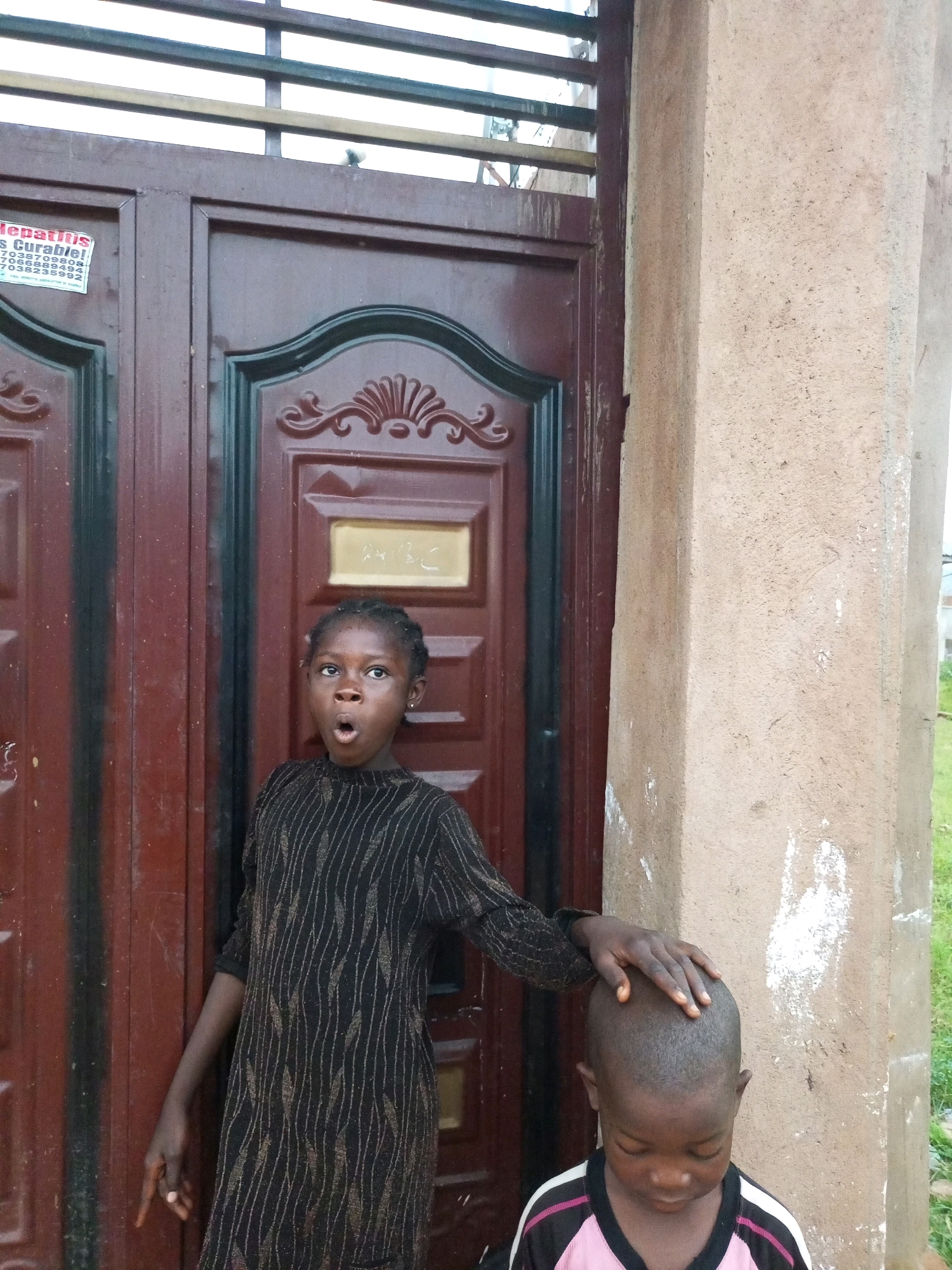 The image shows two young children standing in front of a brown double door with decorative metalwork. The child on the left is a girl with braided hair and is wearing a black dress with a patterned design. She is looking towards the camera with her mouth open in an expression of surprise or excitement. Her left arm is extended outwards, with her hand resting on the head of the child on the right. The child on the right is a boy with a shaved head, wearing a pink and black shirt. He is looking down. The children appear to be outside, with a textured, light-colored concrete pillar to the right of the door. A partially visible sign with text in red and black is attached to the left door. The scene is likely in Tempure, Nigeria, given the context of the prompt.