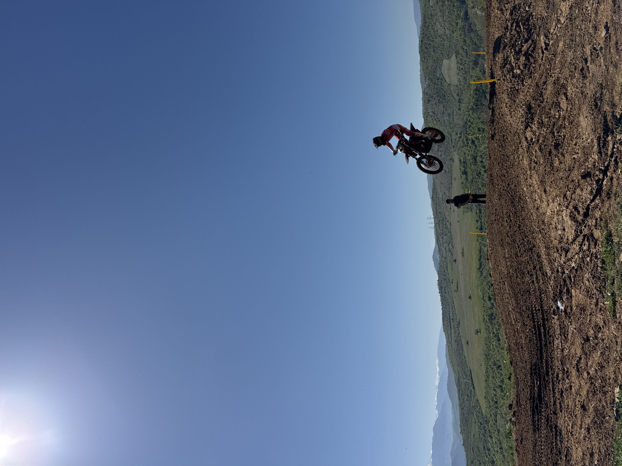 A motocross rider wearing a red suit jumps a dirt bike against a clear blue sky. A second individual stands on the ground near a dirt track, with yellow poles marking the course. Rolling hills and distant mountains are visible in the background. The scene is most likely located in the city of Selishte, Bulgaria, where similar motocross events and terrains are common.